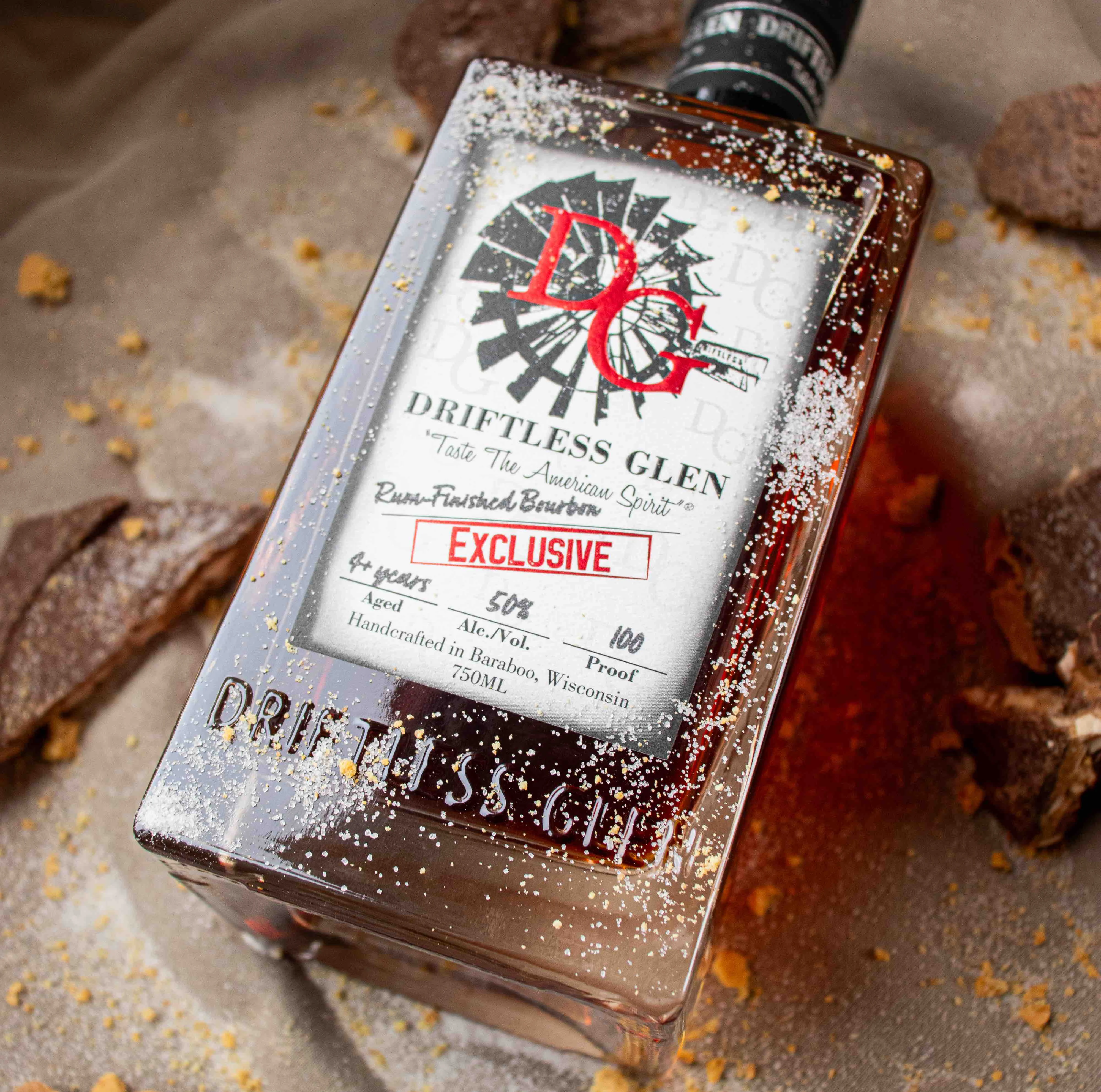 A bottle of Vanilla-Finished Bourbon sit on a whiskey barrel with cinnamon sticks and winter dessert around it.