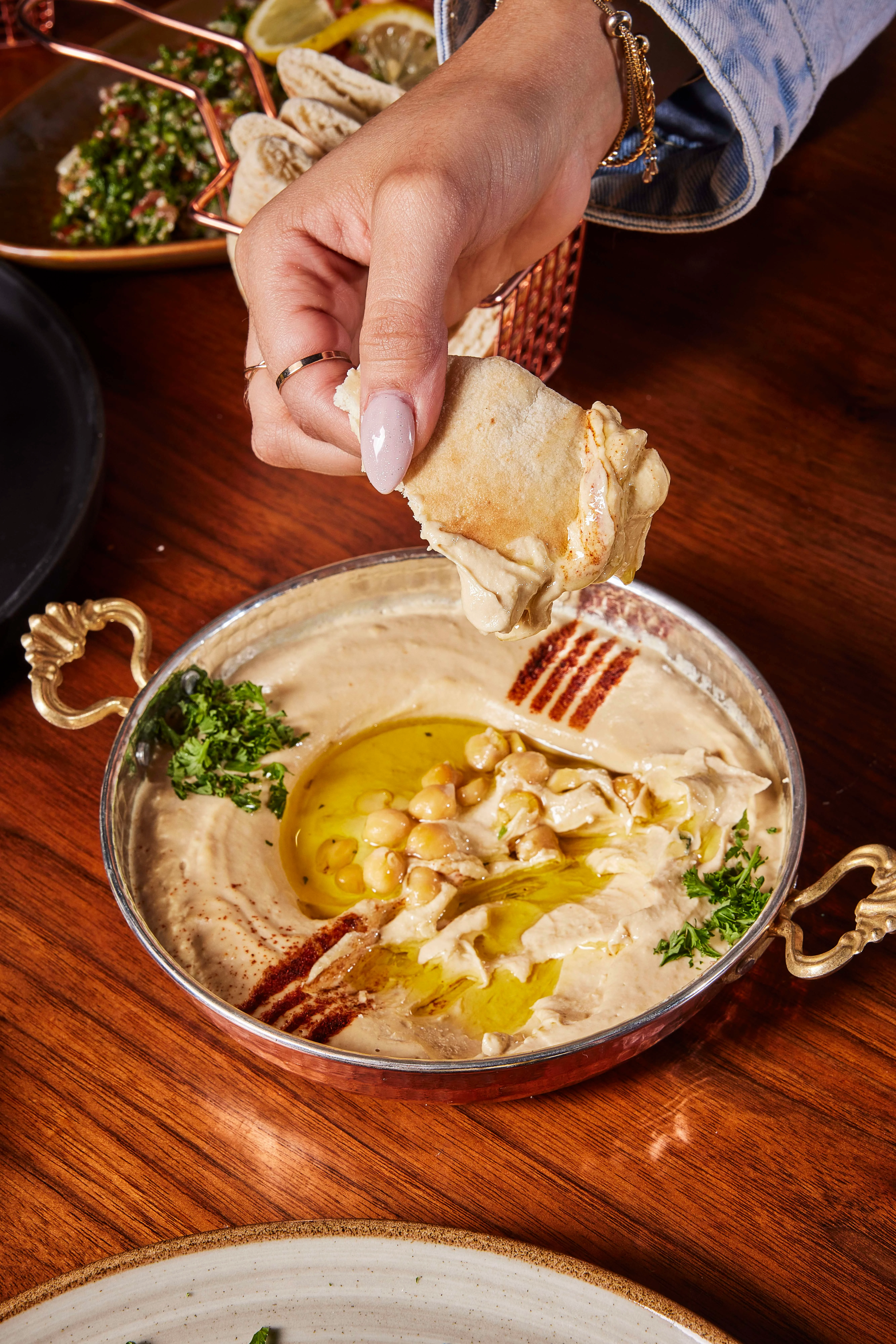 A person dipping a piece of pita bread into a bowl of creamy hummus at Sufrat Grill.