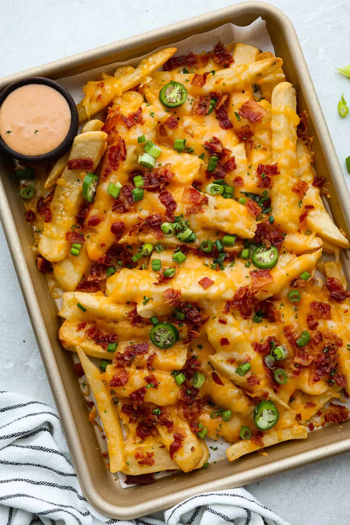 A tray of loaded fries topped with melted cheddar cheese, bacon bits, sliced green onions, and jalapeños, accompanied by a small bowl of dipping sauce.