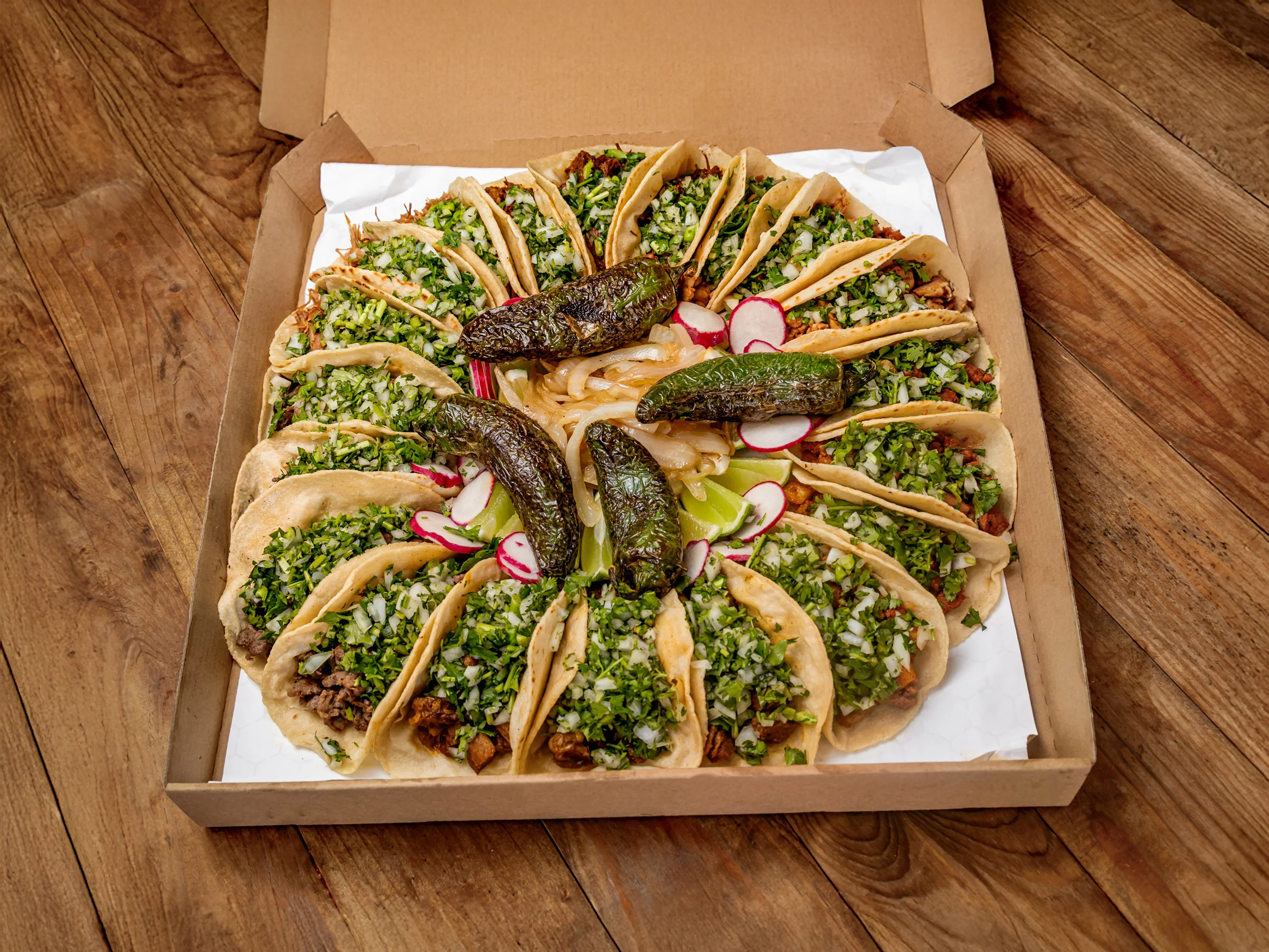 A large assortment of tacos arranged in a box, filled with meat, onions, and various toppings, garnished with jalapeños and radishes.