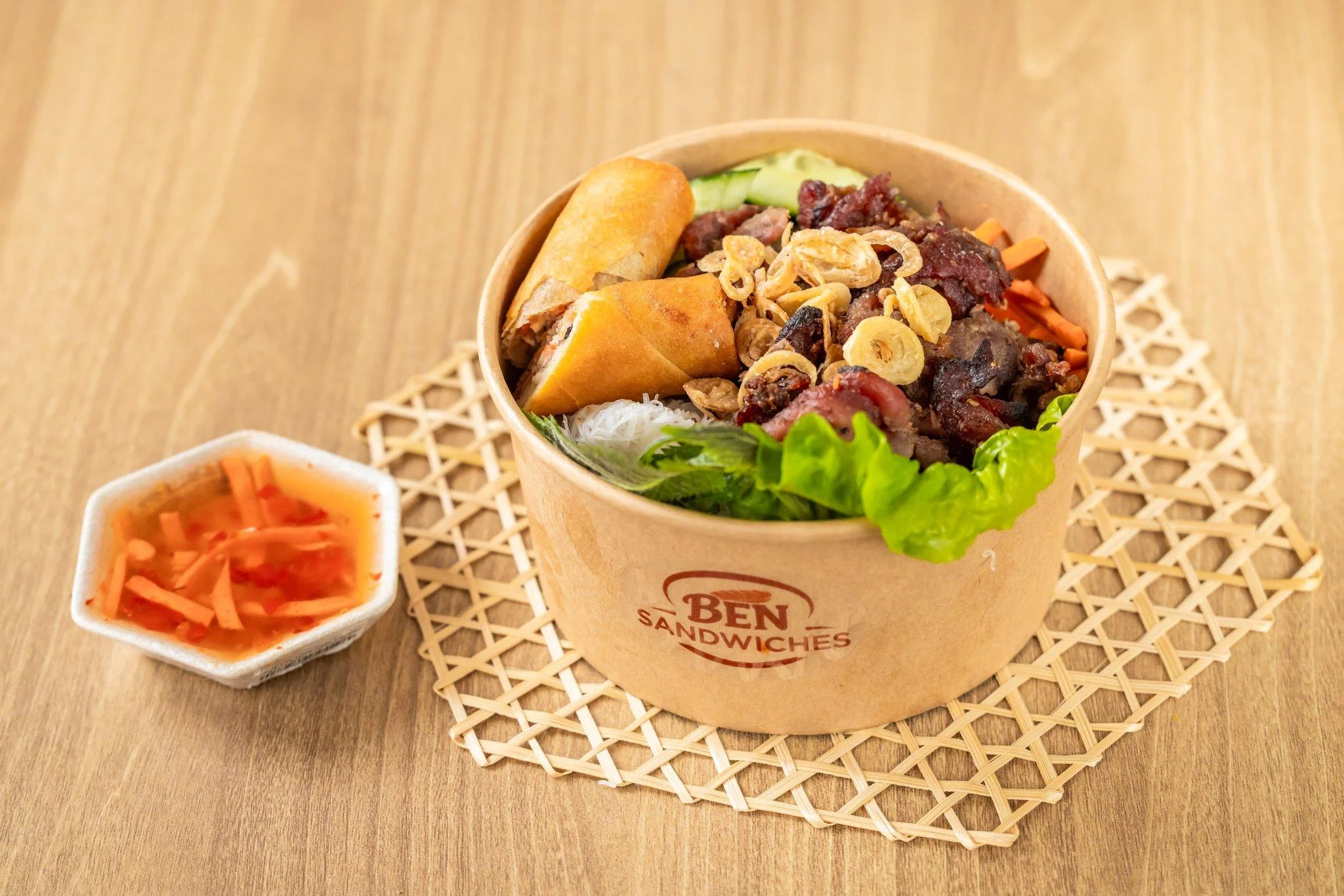 A bowl featuring a variety of ingredients including fried spring rolls, meat, and vegetables. Accompanied by a small dish of dipping sauce.
