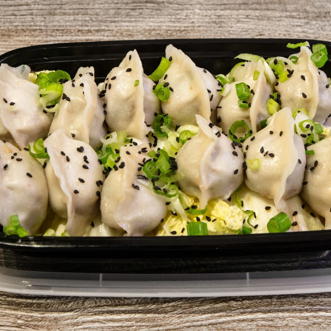 Steamed dumplings topped with green onions and sesame seeds in a black container.