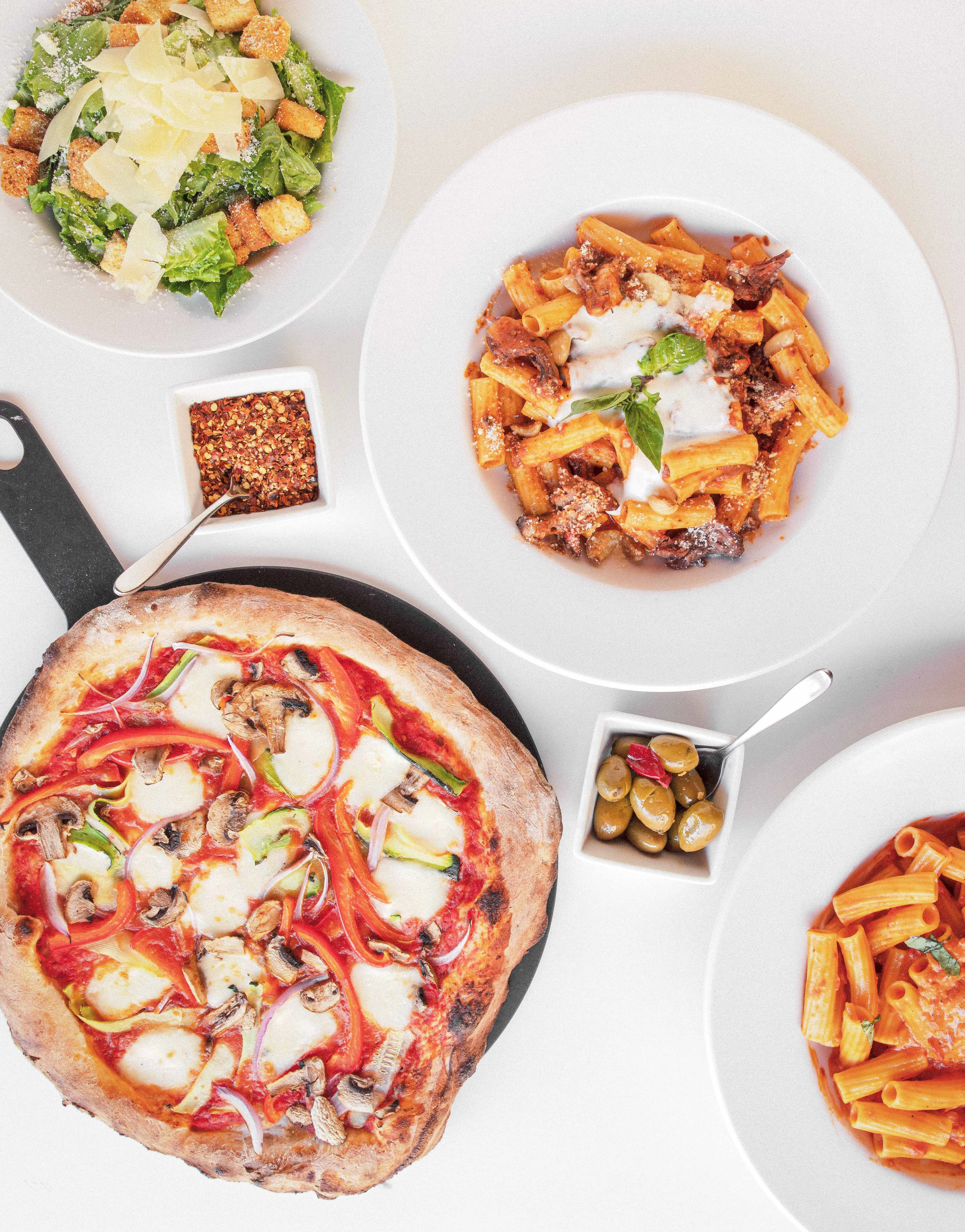 various assortment of brick oven pizza, fresh pasta, and salads on a blank white table. A side of peppers and olives in between the dishes