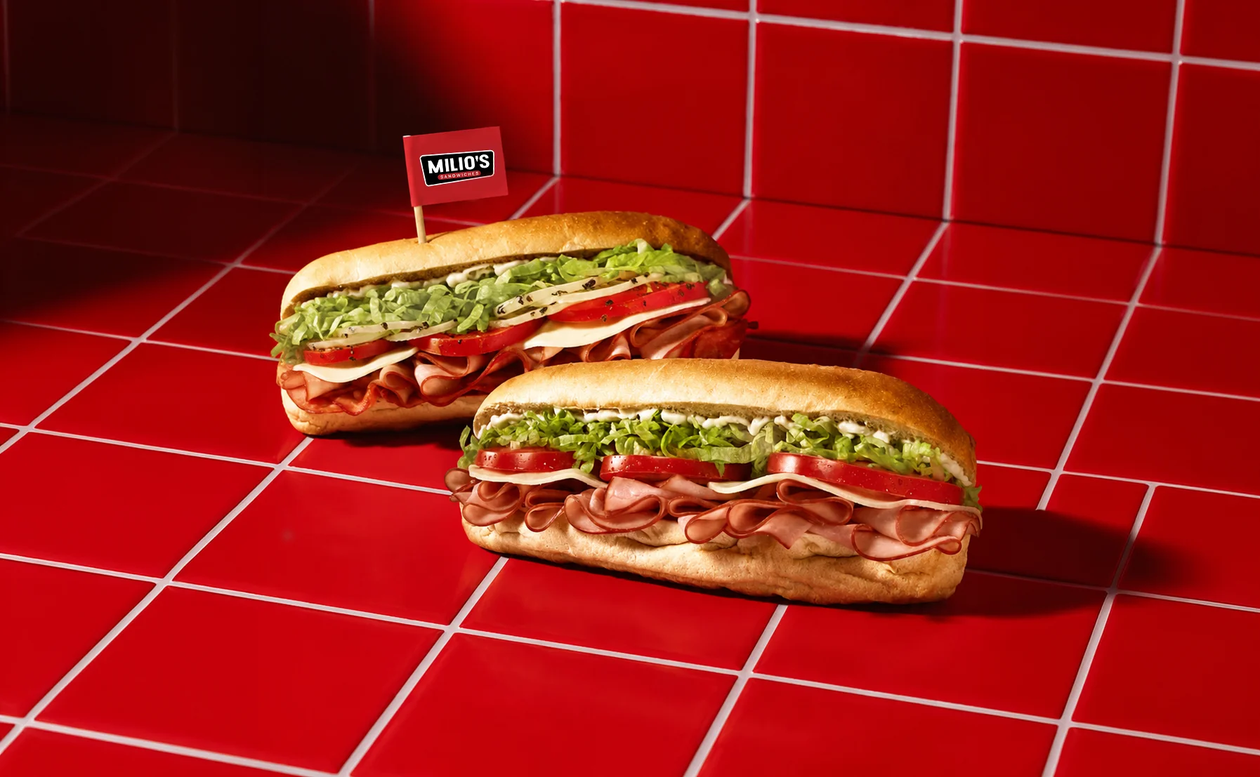 Two Milio's sub sandwiches on a red tile backdrop.