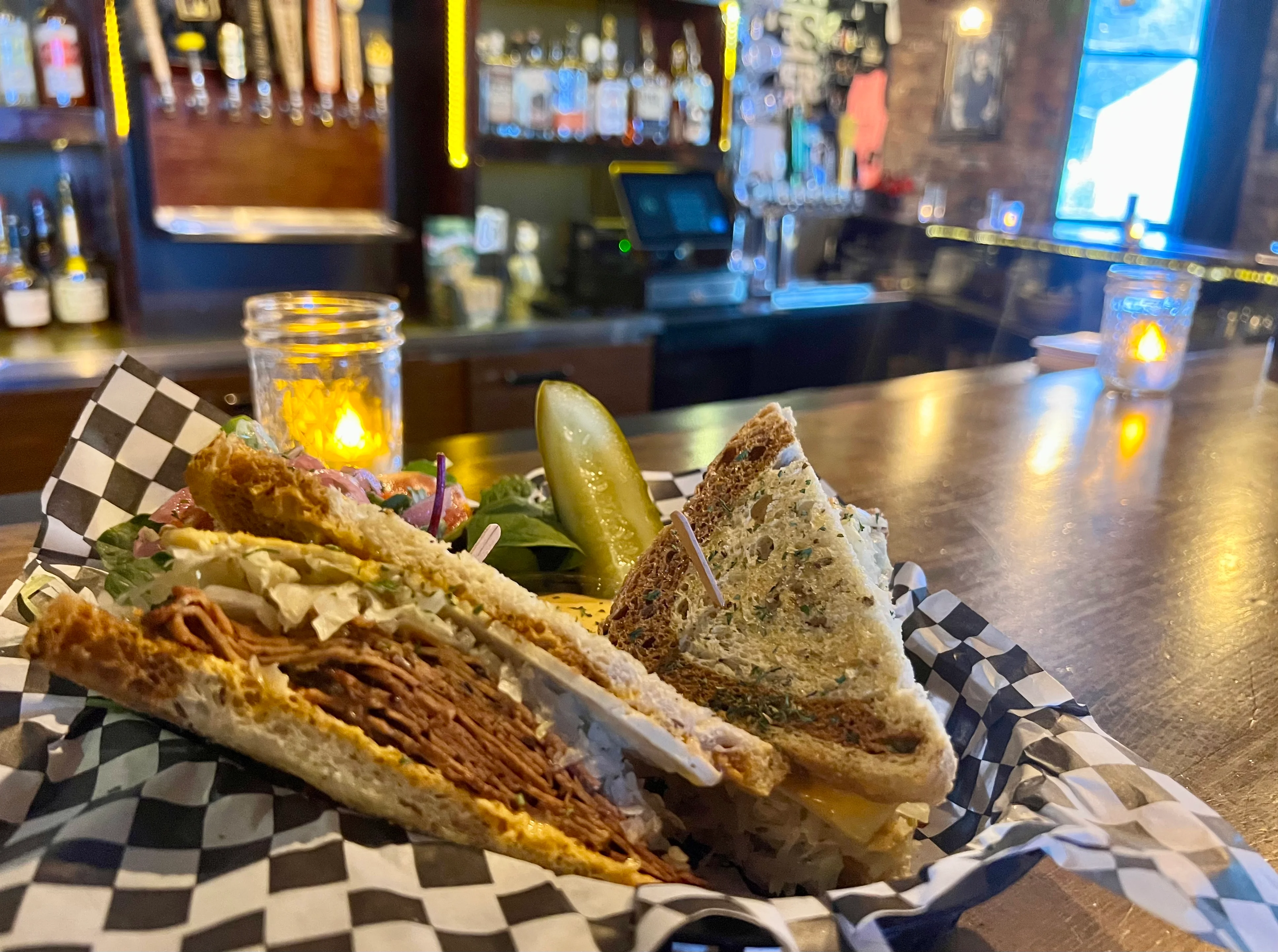 A juicy hamburger with lettuce, tomato, and cheese served in a checkered paper basket alongside a pickle.
