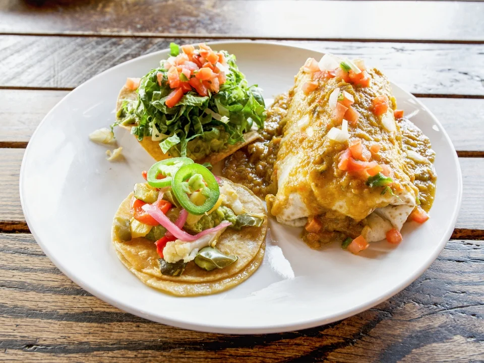 A plate featuring a burrito topped with green sauce and garnished with diced tomatoes, alongside a taco filled with vegetables and a salad of shredded lettuce and tomatoes. The food is served on a white plate against a wooden table.