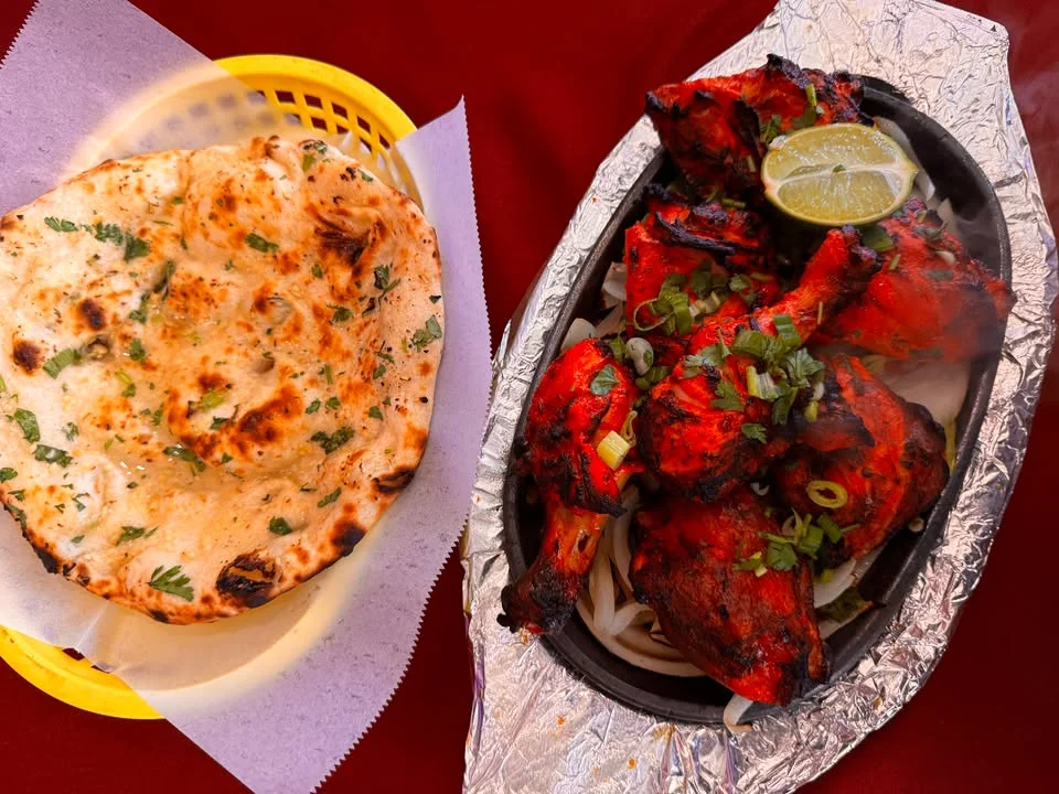 A sizzling platter of grilled chicken, bright red in color, served with a piece of naan bread and garnished with herbs.
