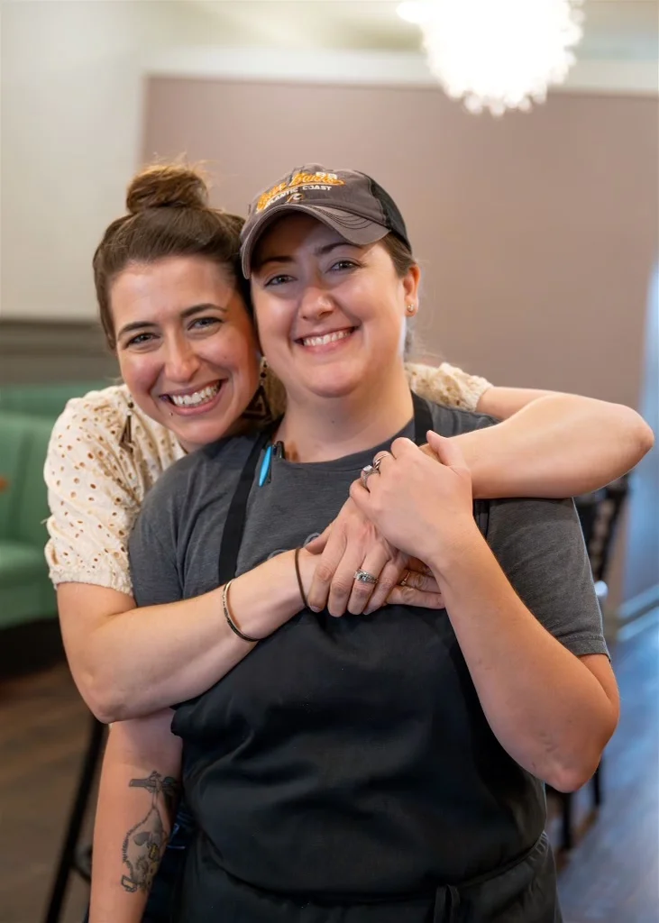 Two smiling restaurant staff members embracing in a cozy dining space