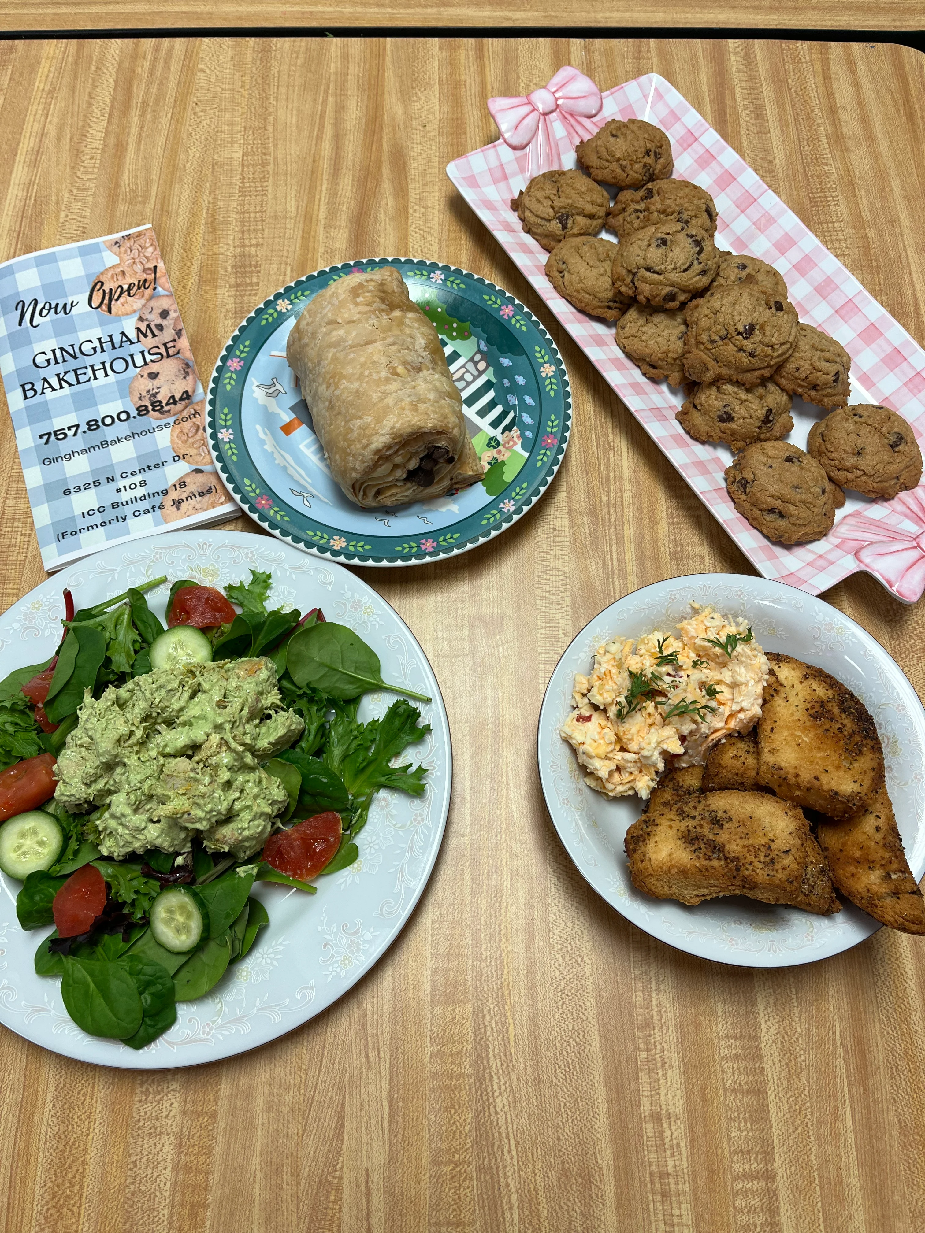 A table with plates of food: chocolate chip cookies, a chocolate croissant, green goddess chicken salad on fresh greens, and pimento cheese with homemade herb brioche crackers.