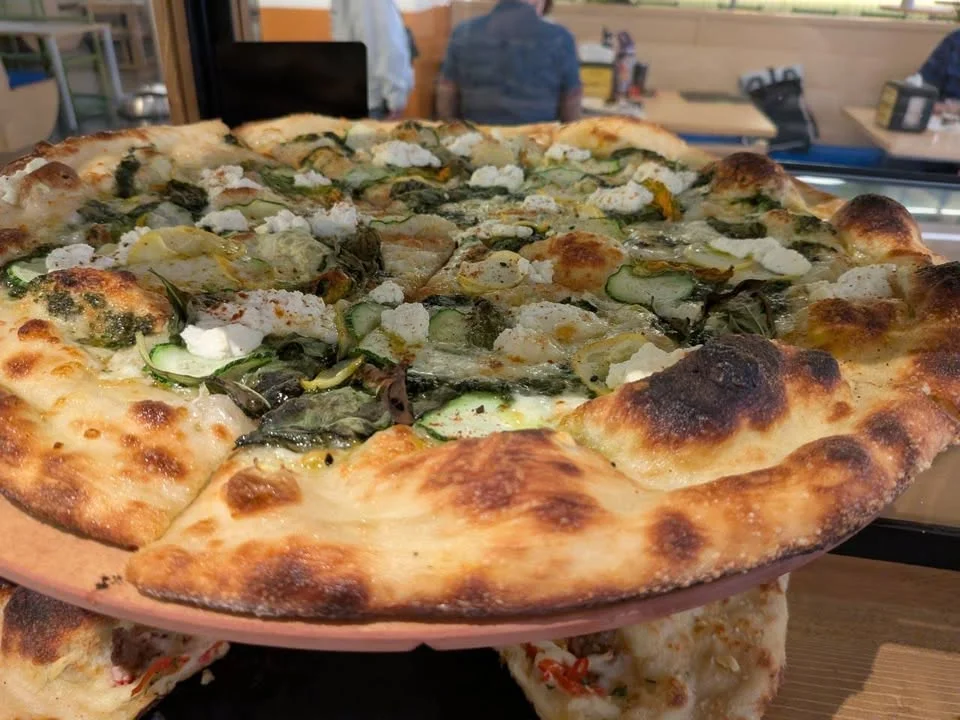 A vegetable pizza topped with greens and slices of zucchini, served on a wooden tray.