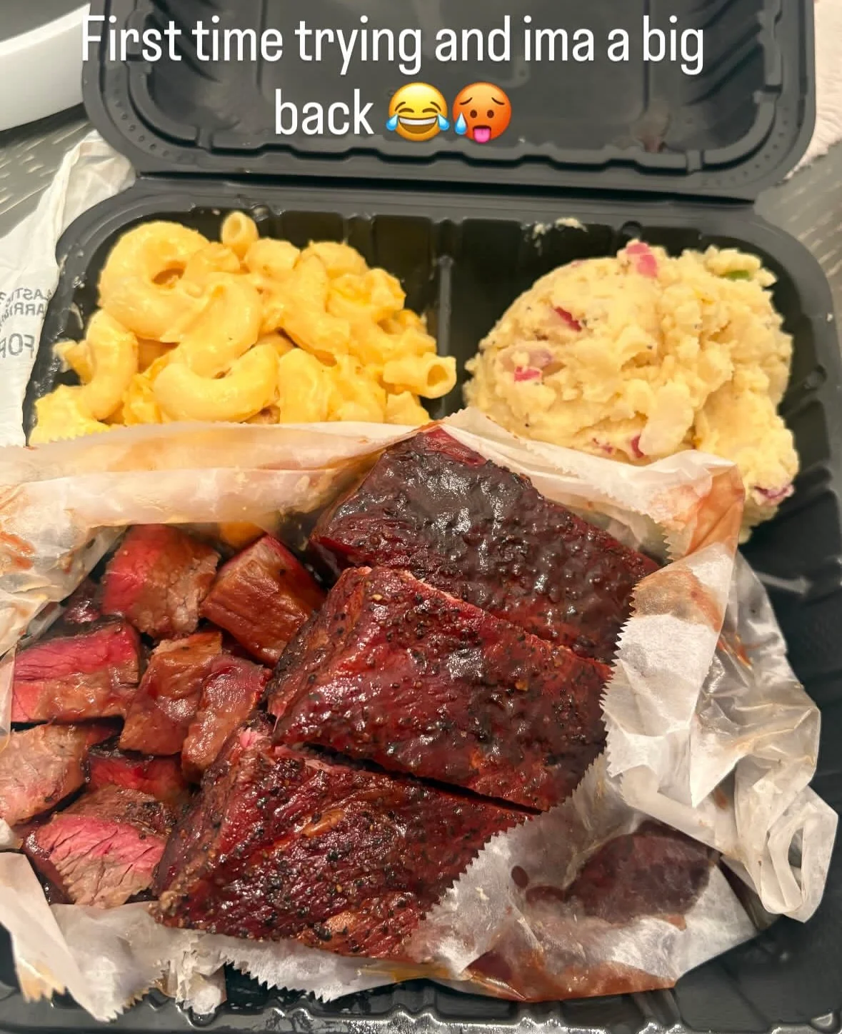 A takeout container with barbecue beef, macaroni and cheese, and a potato salad, featuring assorted cuts of meat alongside side dishes.