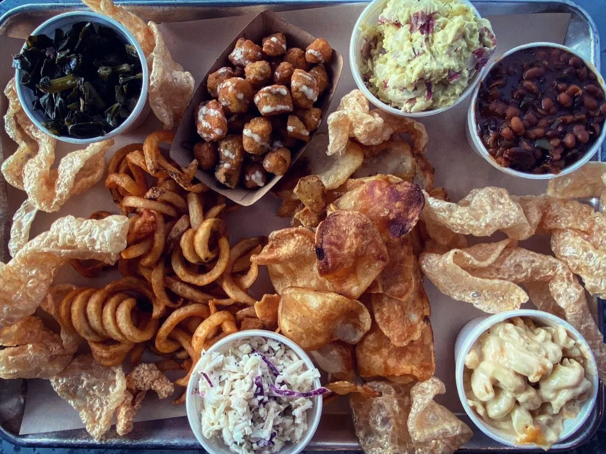 A platter filled with various side dishes including crispy pork rinds, curly fries, and multiple cups of salads and beans, arranged neatly in a tray.