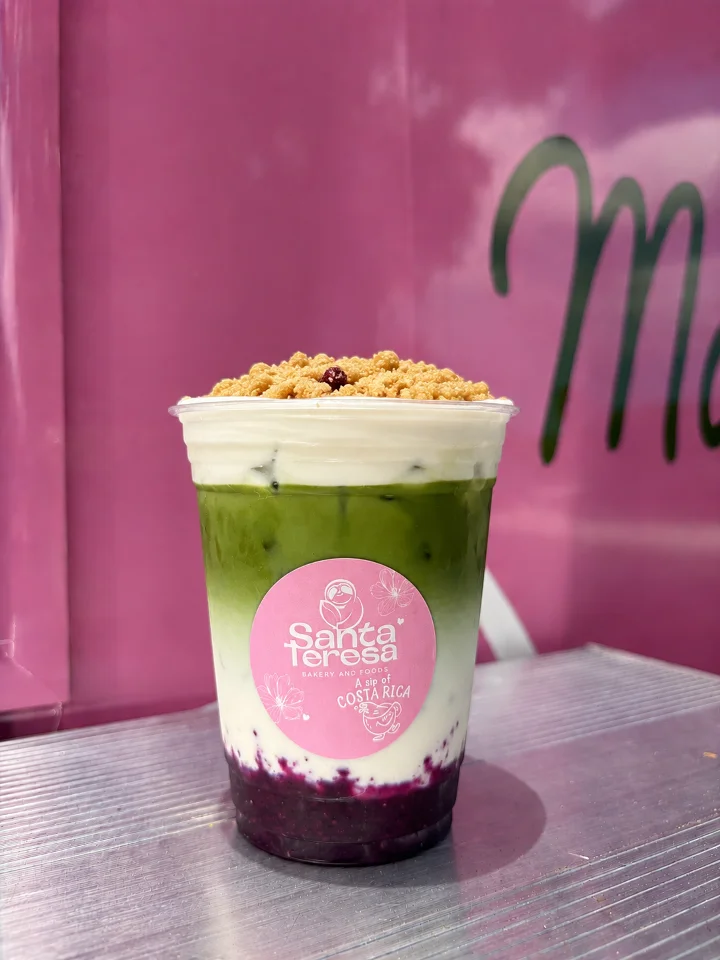 A colorful layered drink in a clear cup featuring green matcha, white cream, and purple berry layers topped with a crunchy crumb.