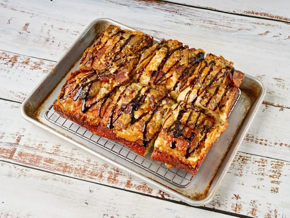 A rectangular tray of dessert pizza topped with chocolate drizzle and various toppings, showcasing a golden-brown crust.