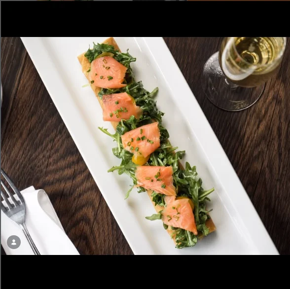 A long, narrow plate featuring a bed of arugula topped with slices of smoked salmon and garnished with herbs, alongside a glass of sparkling wine.