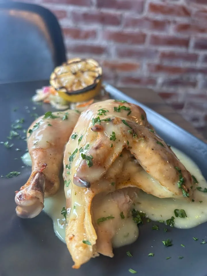 A plated dish of two tender chicken pieces covered in a creamy sauce, garnished with herbs, alongside a charred lemon.