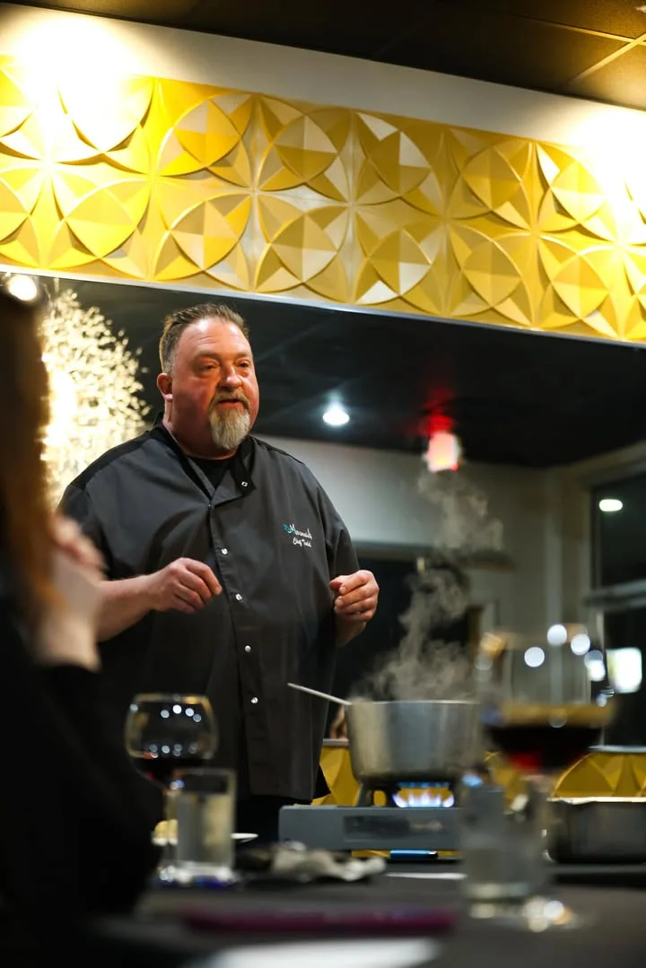 Chef Todd Golden stands at the front of a dining room, mid-demonstration, wearing a black chef coat with his name embroidered on it. Steam rises from a pot on a portable burner in front of him, while guests watch attentively, glasses of wine and water on the table. A modern gold geometric wall design and ambient lighting create a warm, upscale atmosphere.