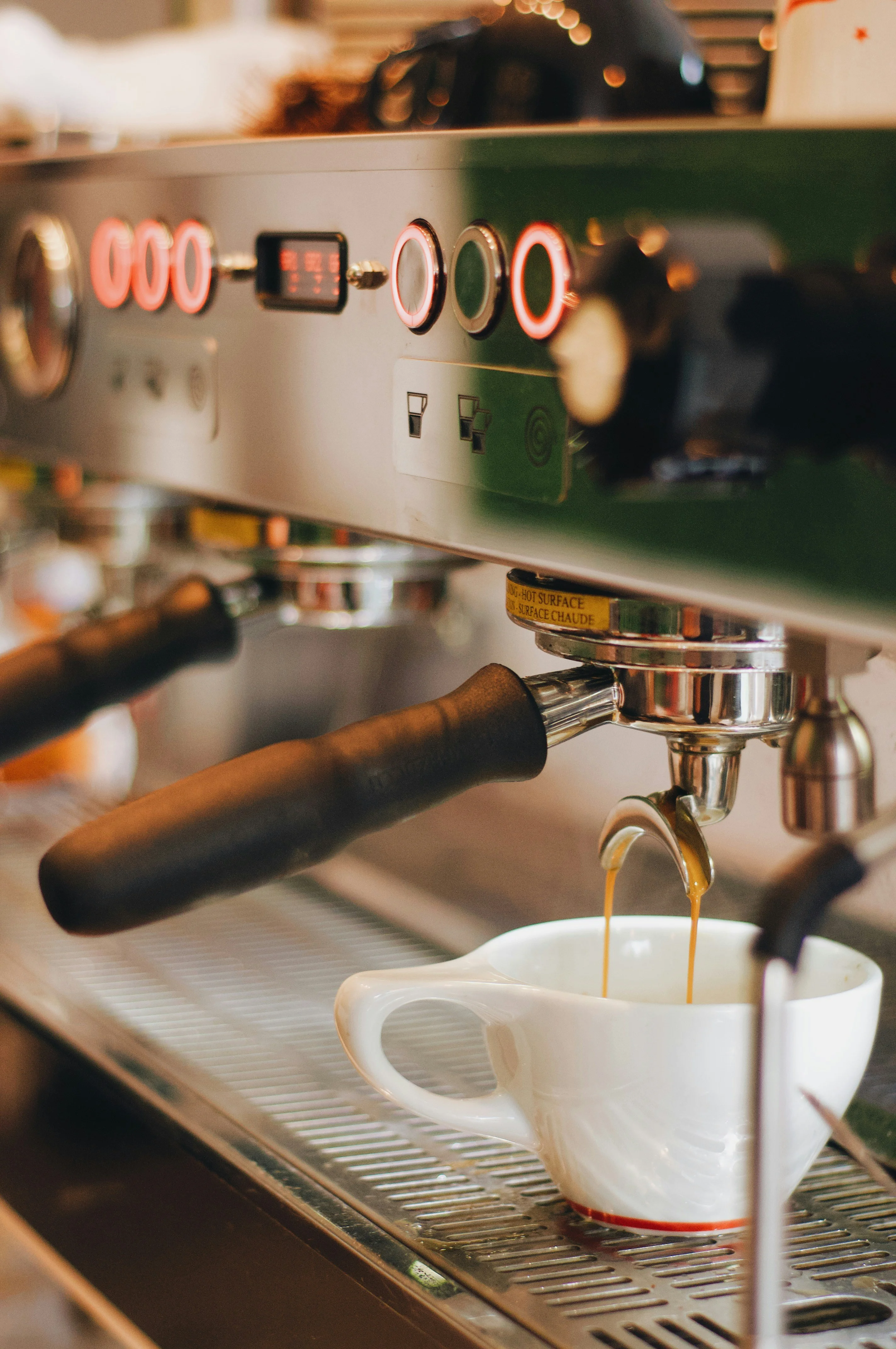 A hand holding a glass of latte art coffee in front of large green leaves, showcasing a decorative milk pattern.