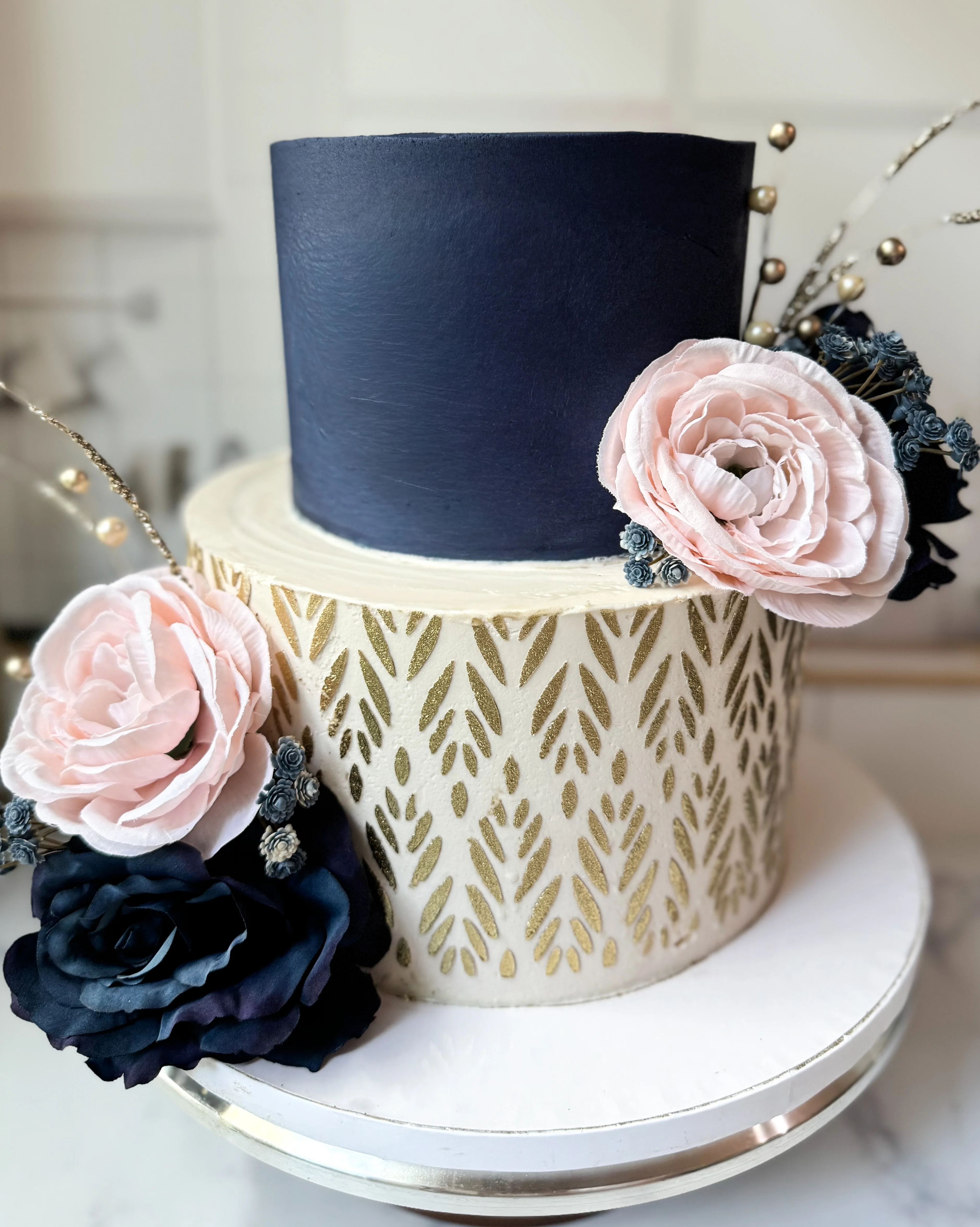 A two-tier cake with a dark blue top tier and a patterned gold and cream bottom tier, adorned with pink and navy blue flowers.