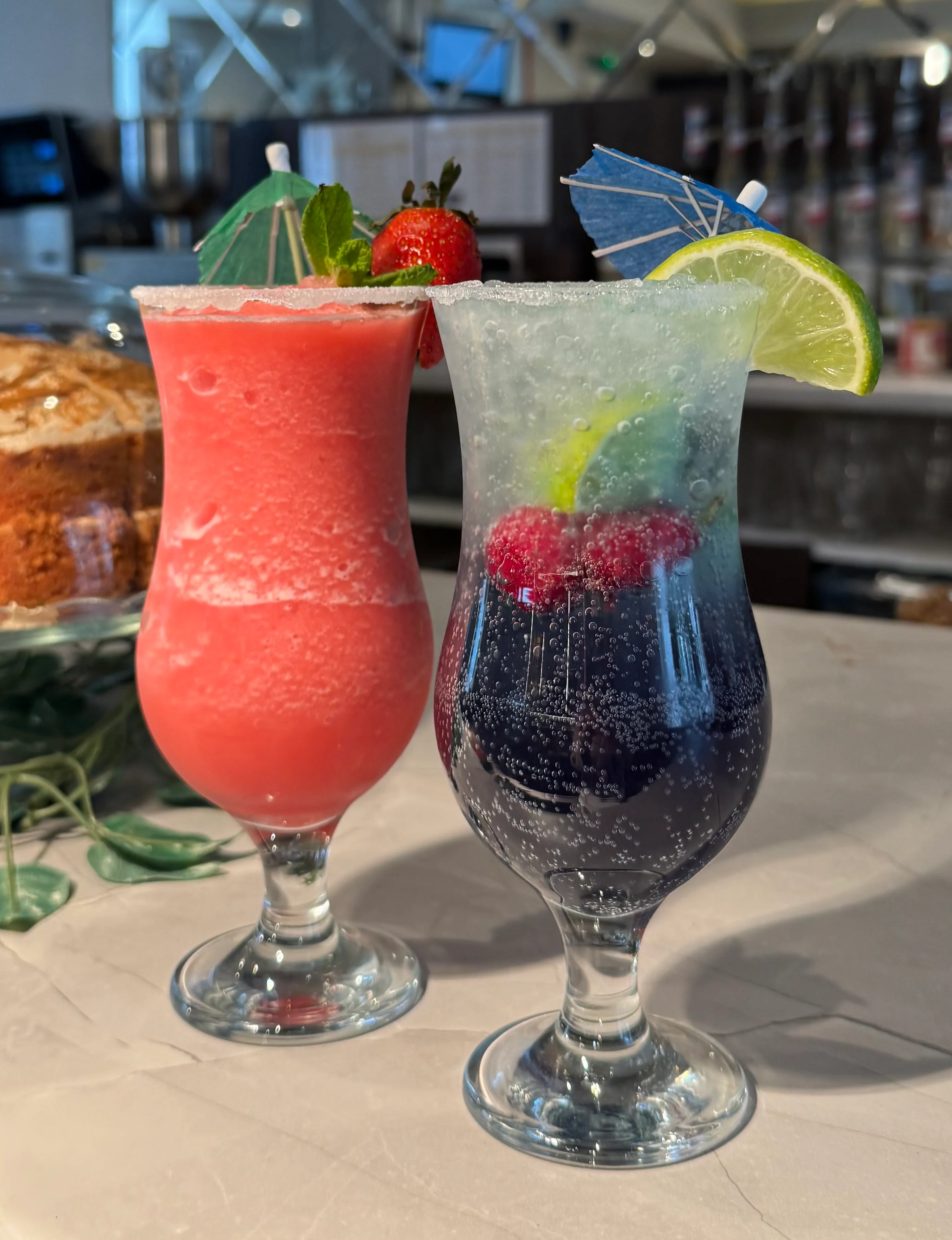 Two colorful cocktails with garnishes on a table in a restaurant.