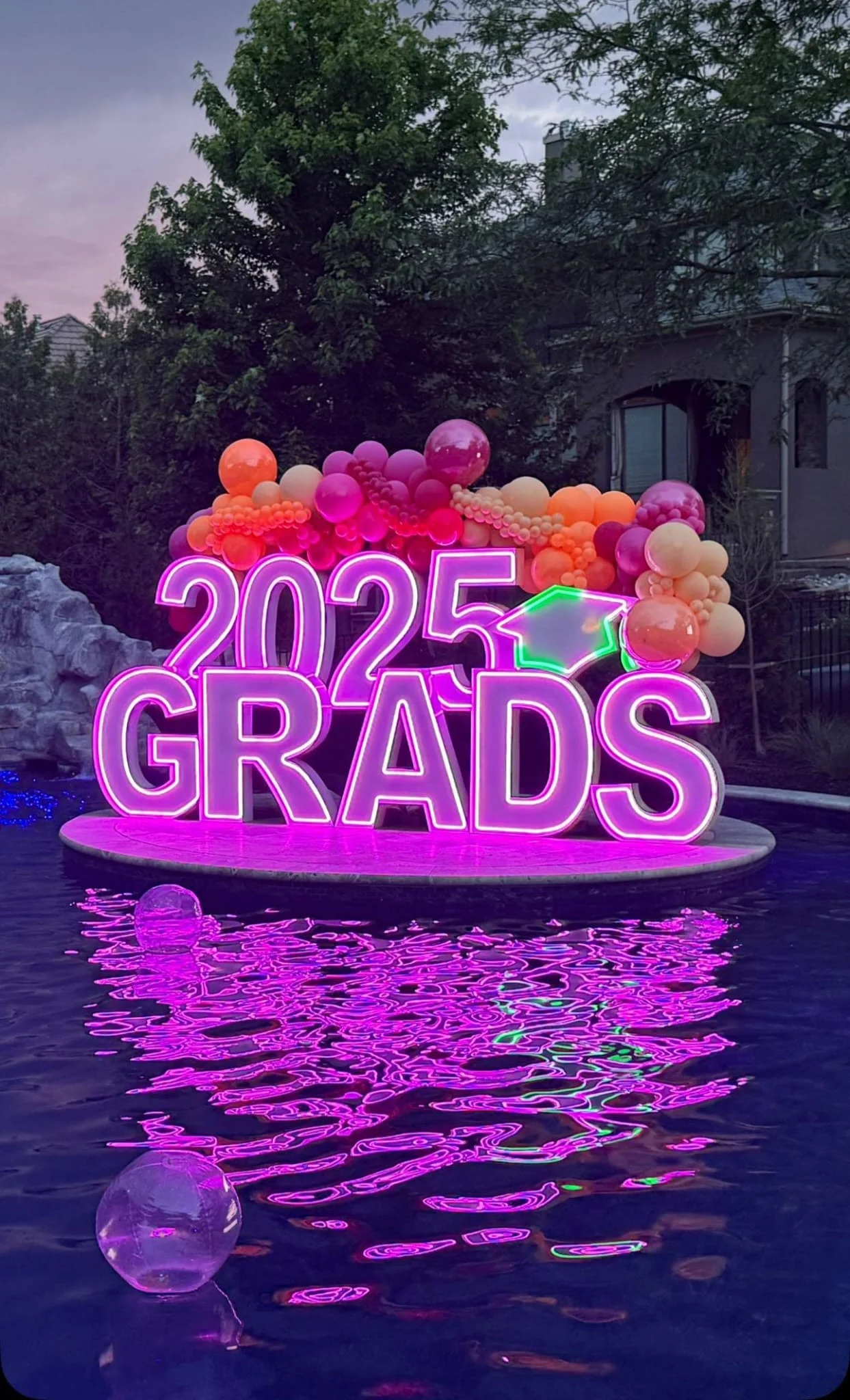A vibrant display for a graduation celebration featuring large neon letters '2025 GRADS' surrounded by colorful balloons, set near a pool.