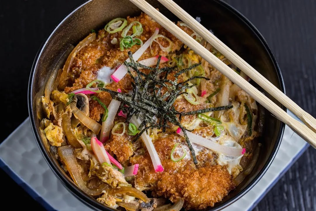 A bowl of katsu don featuring breaded pork cutlet, egg, and onions topped with green onions and nori.