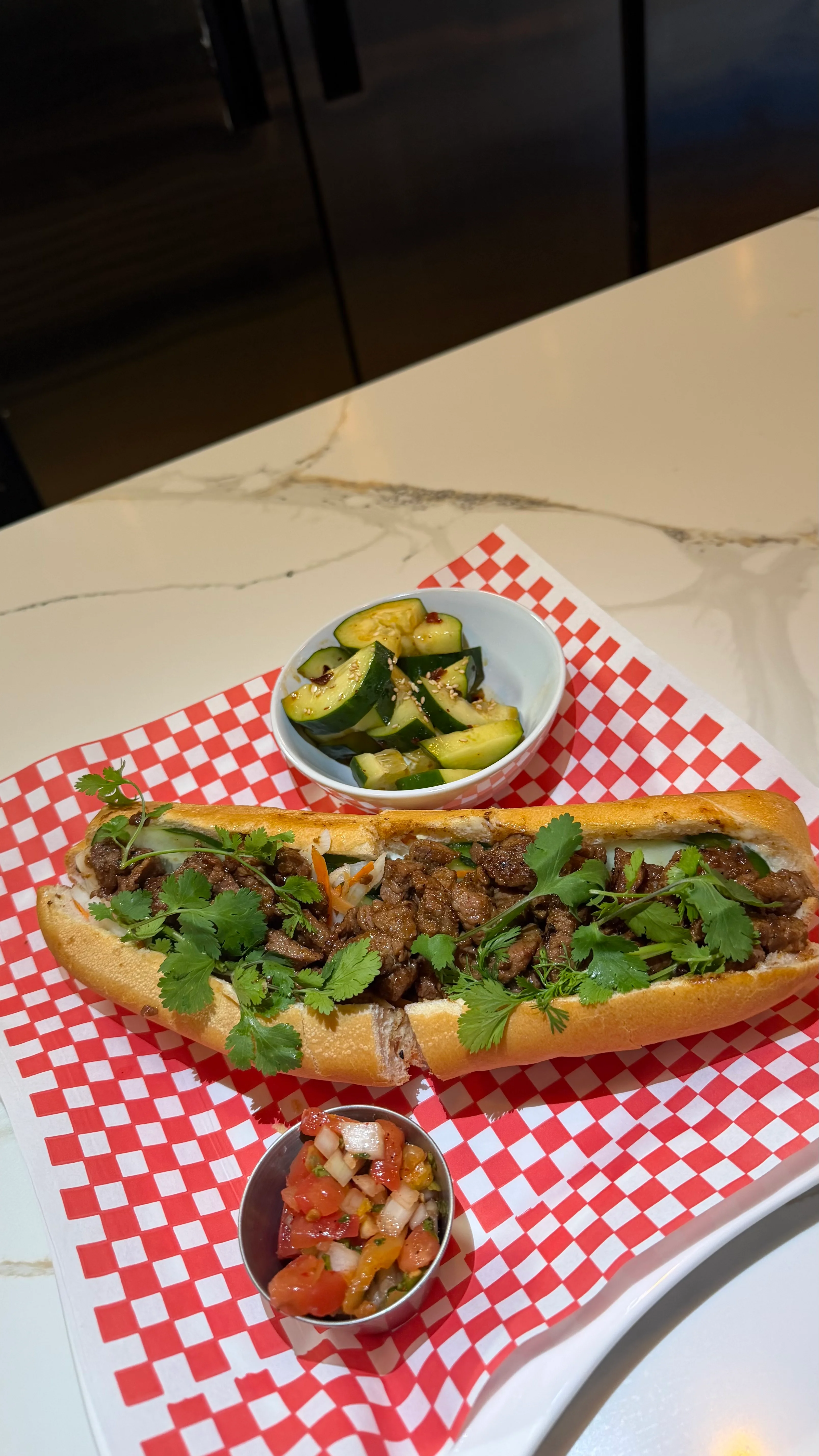 A banh mi sandwich filled with beef, cucumbers, and cilantro, served with a side of fresh salsa and cucumber salad.
