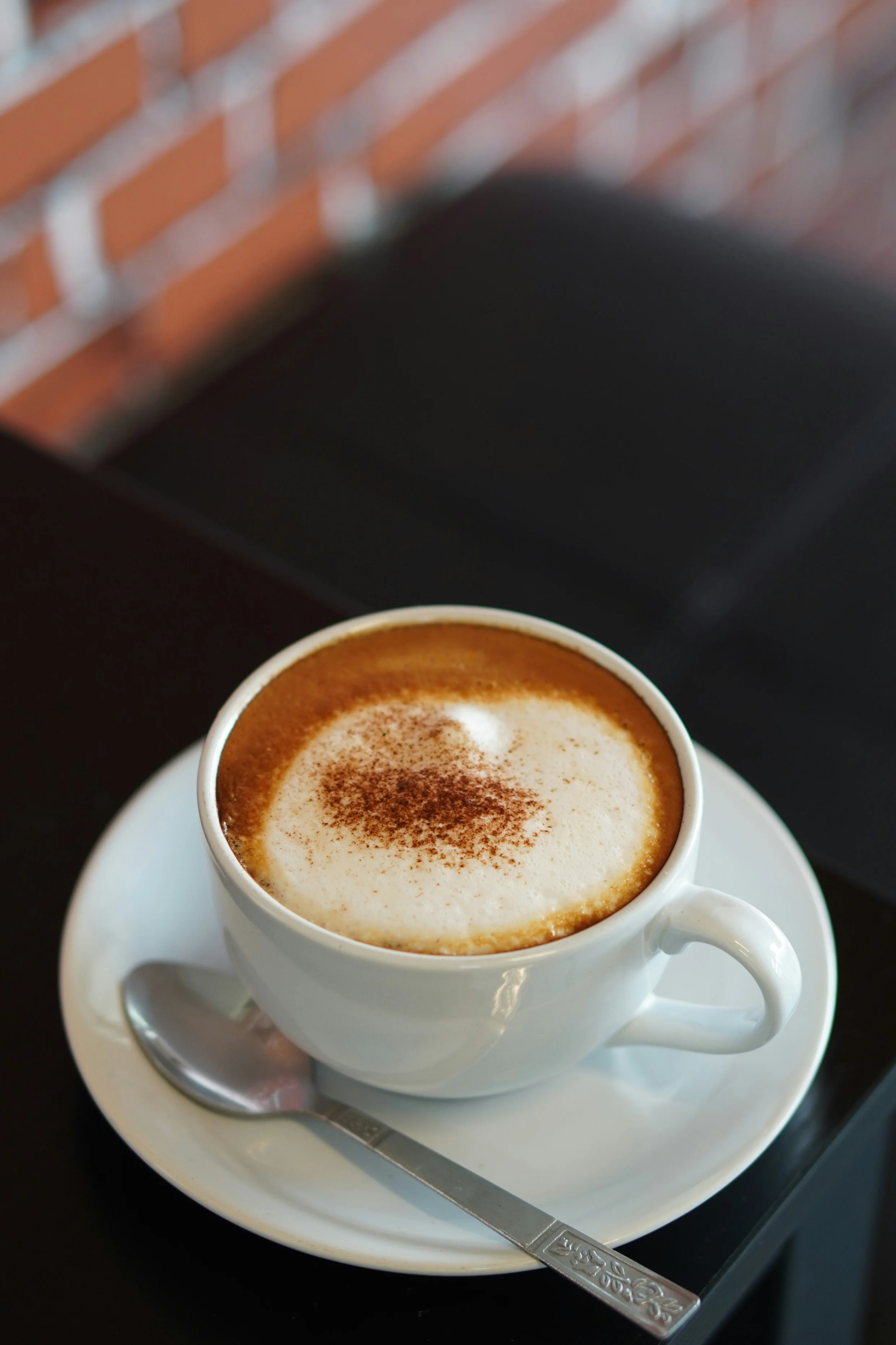 A cup of cappuccino topped with frothy milk and sprinkled with cocoa powder, placed on a white saucer with a spoon next to it.