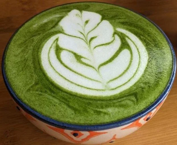 A person whisking a green tea mixture in a traditional bowl using a bamboo whisk. The vibrant green color of the tea is visible in the bowl.