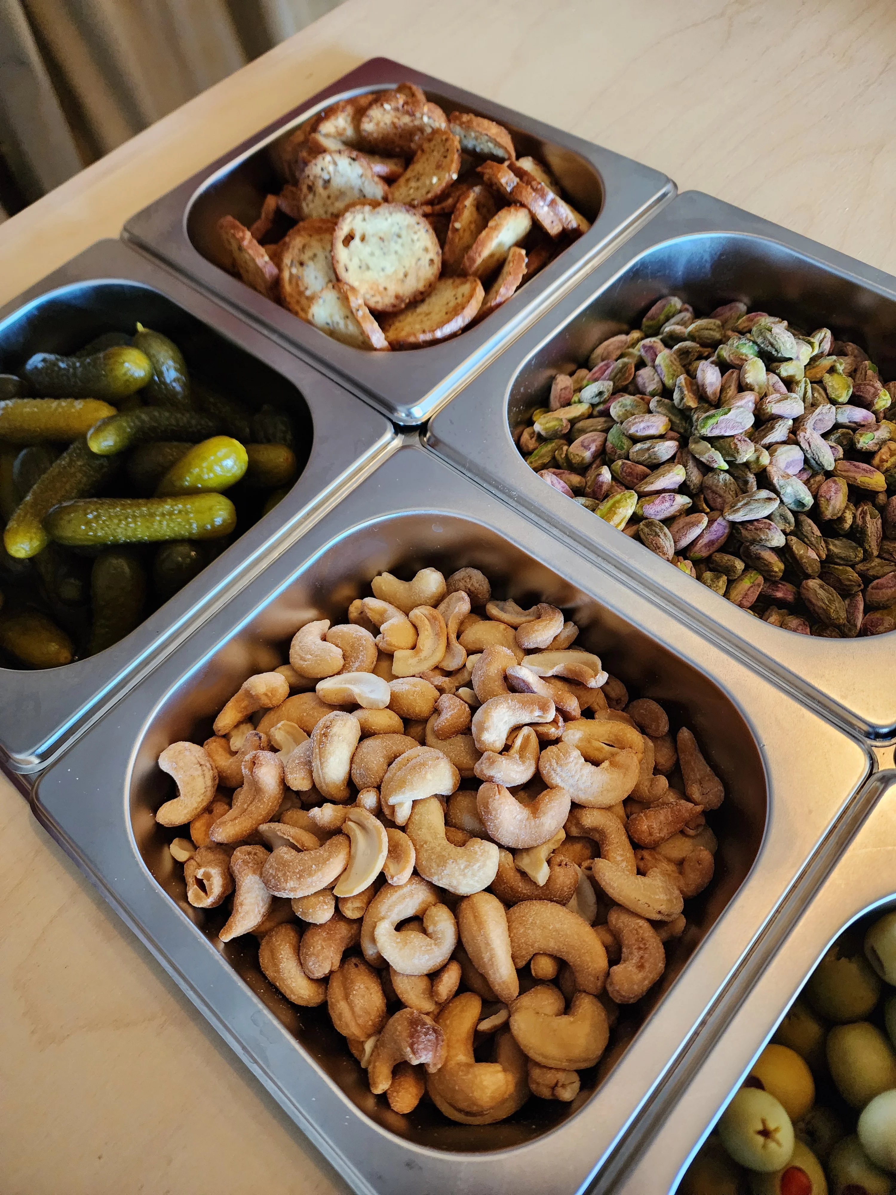 An array of snacks in metal containers, including pickles, nuts, and chips.