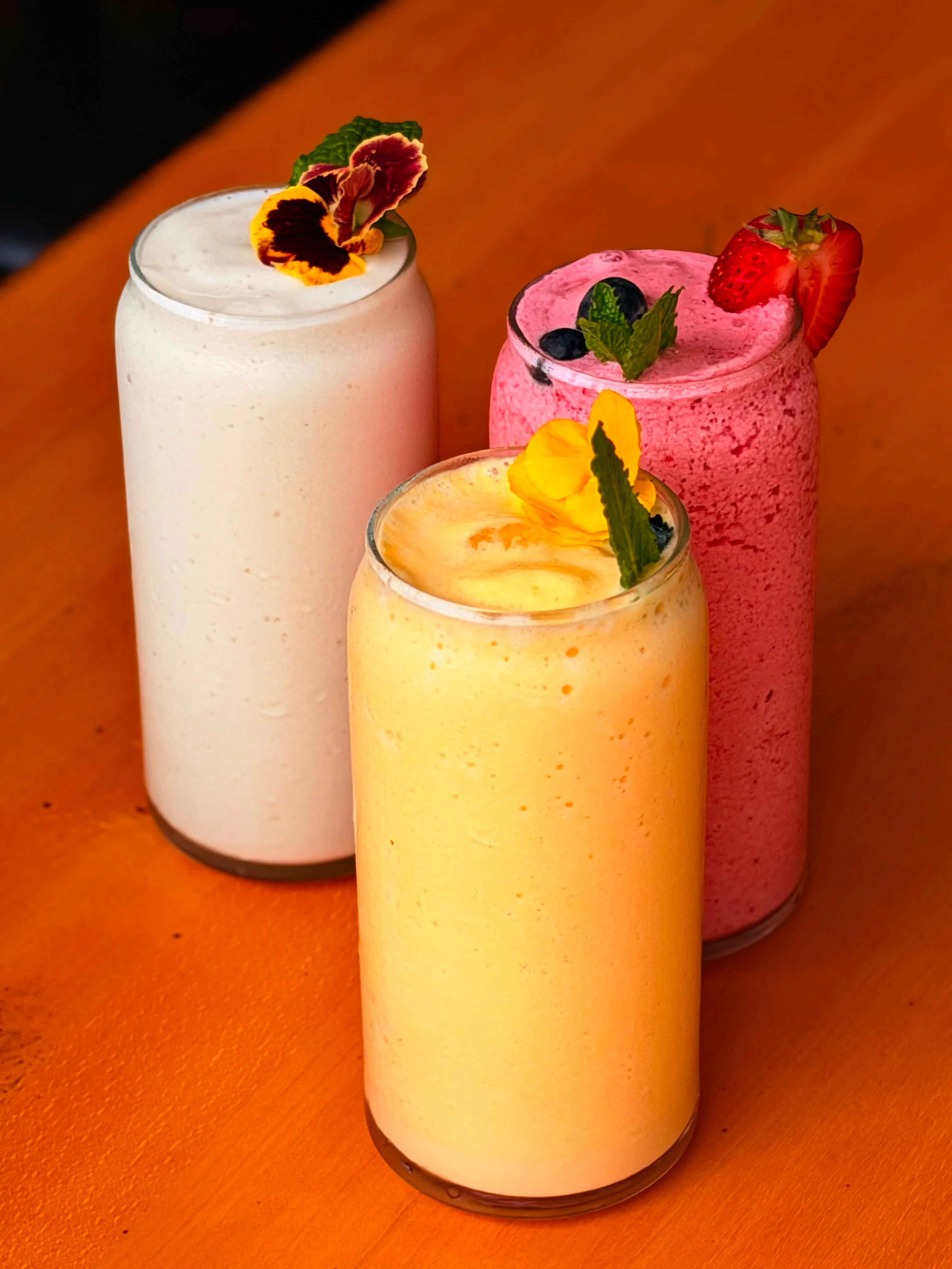 Three smoothies in glass cups, colored pink, yellow, and white, each garnished with fruit and mint leaves.