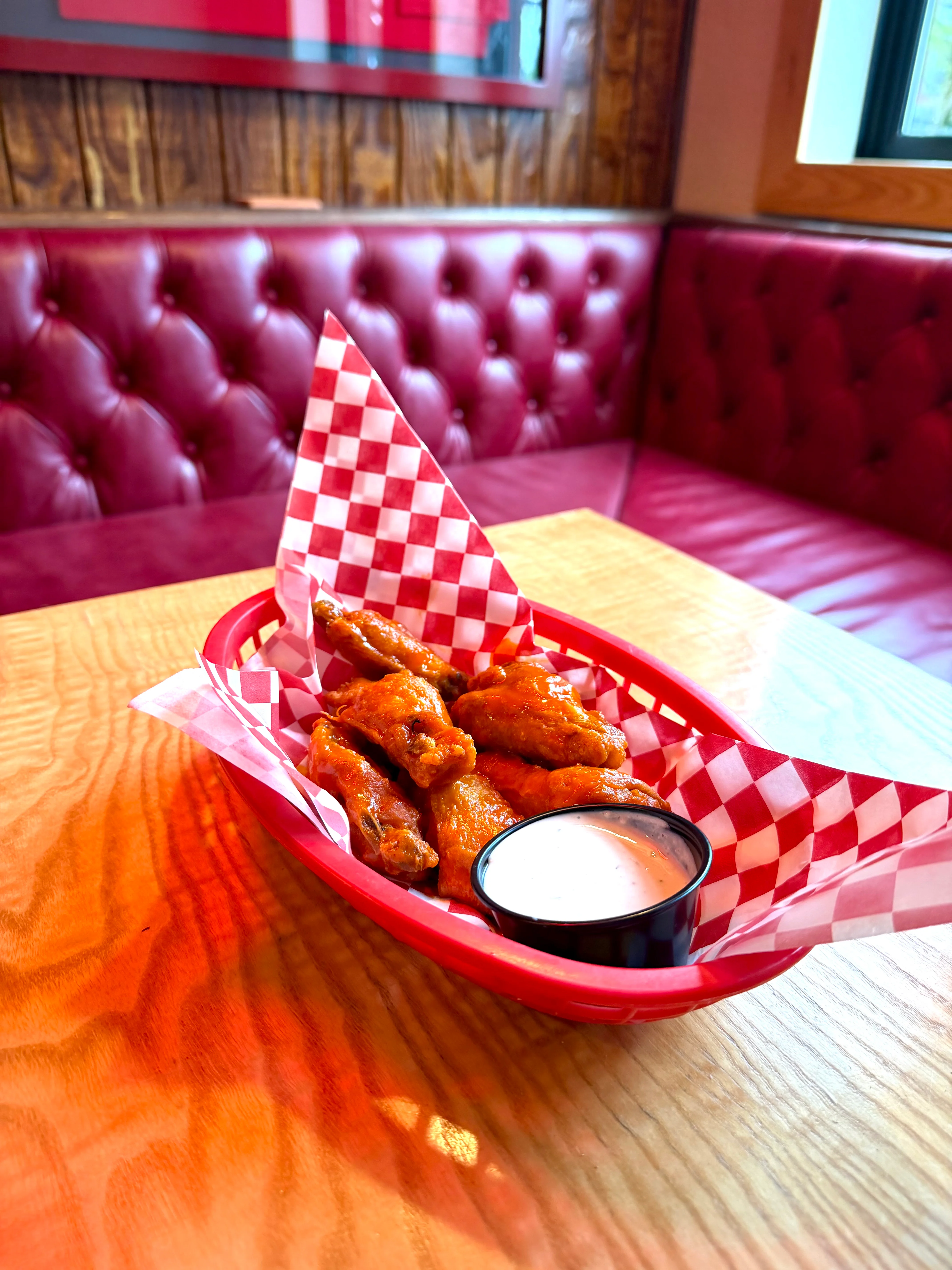 A serving of fried chicken wings presented in a basket with checkered paper, accompanied by a small cup of dipping sauce.