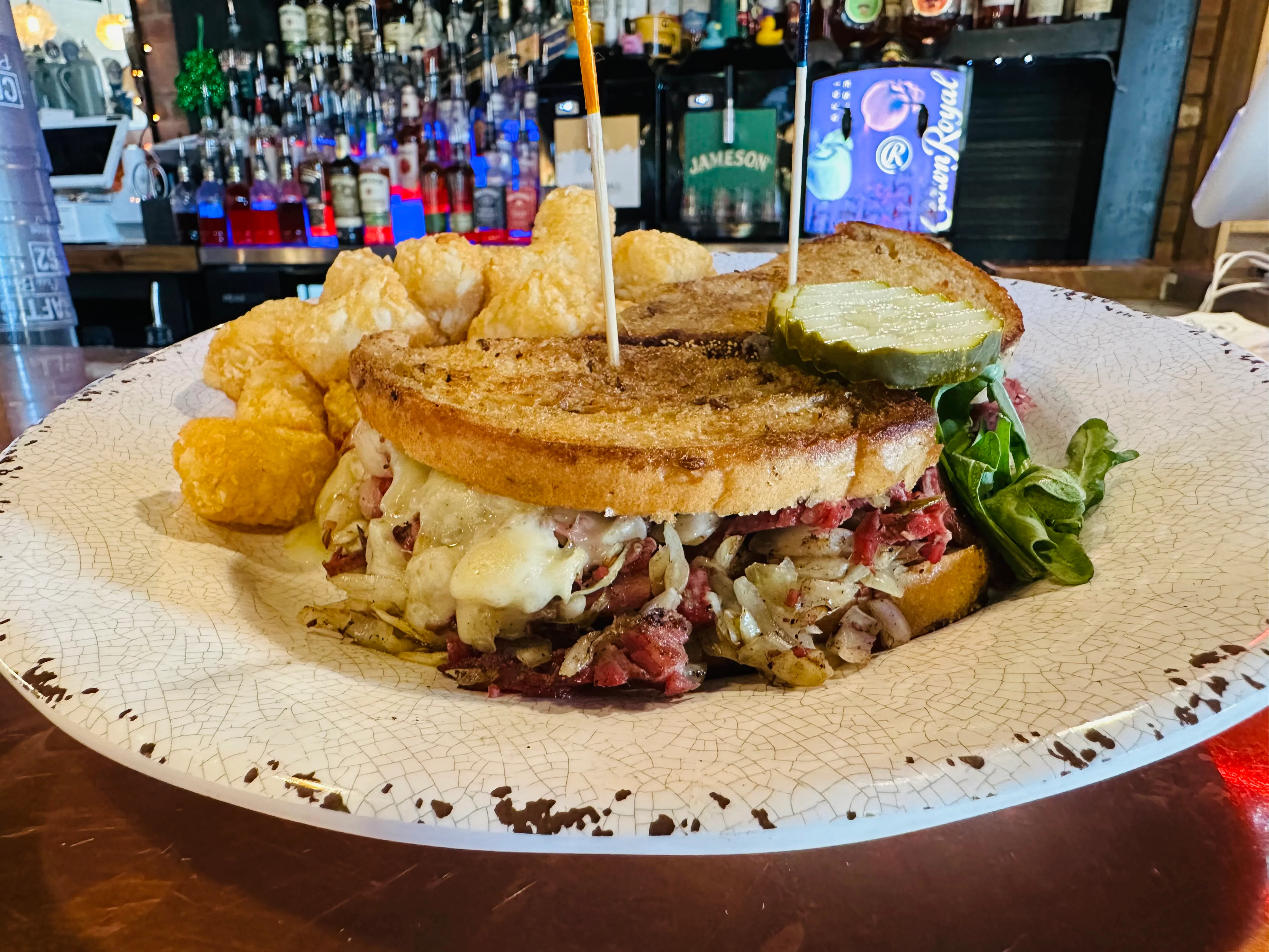 A hearty sandwich featuring layers of meat, cheese, and onions, accompanied by crunchy potato bites and a pickle slice, served on a decorative plate.