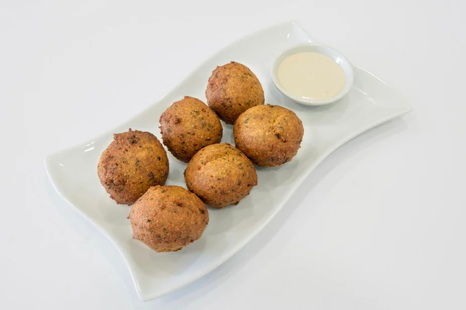 A plate of six crispy falafel balls served with a small dish of dipping sauce. The falafel are golden brown and have a crunchy texture.