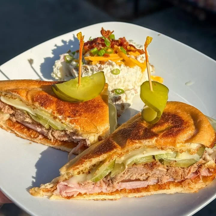 A toasted sandwich filled with layers of meat and pickles, served on a plate with a side of potato salad topped with bacon and cheese.