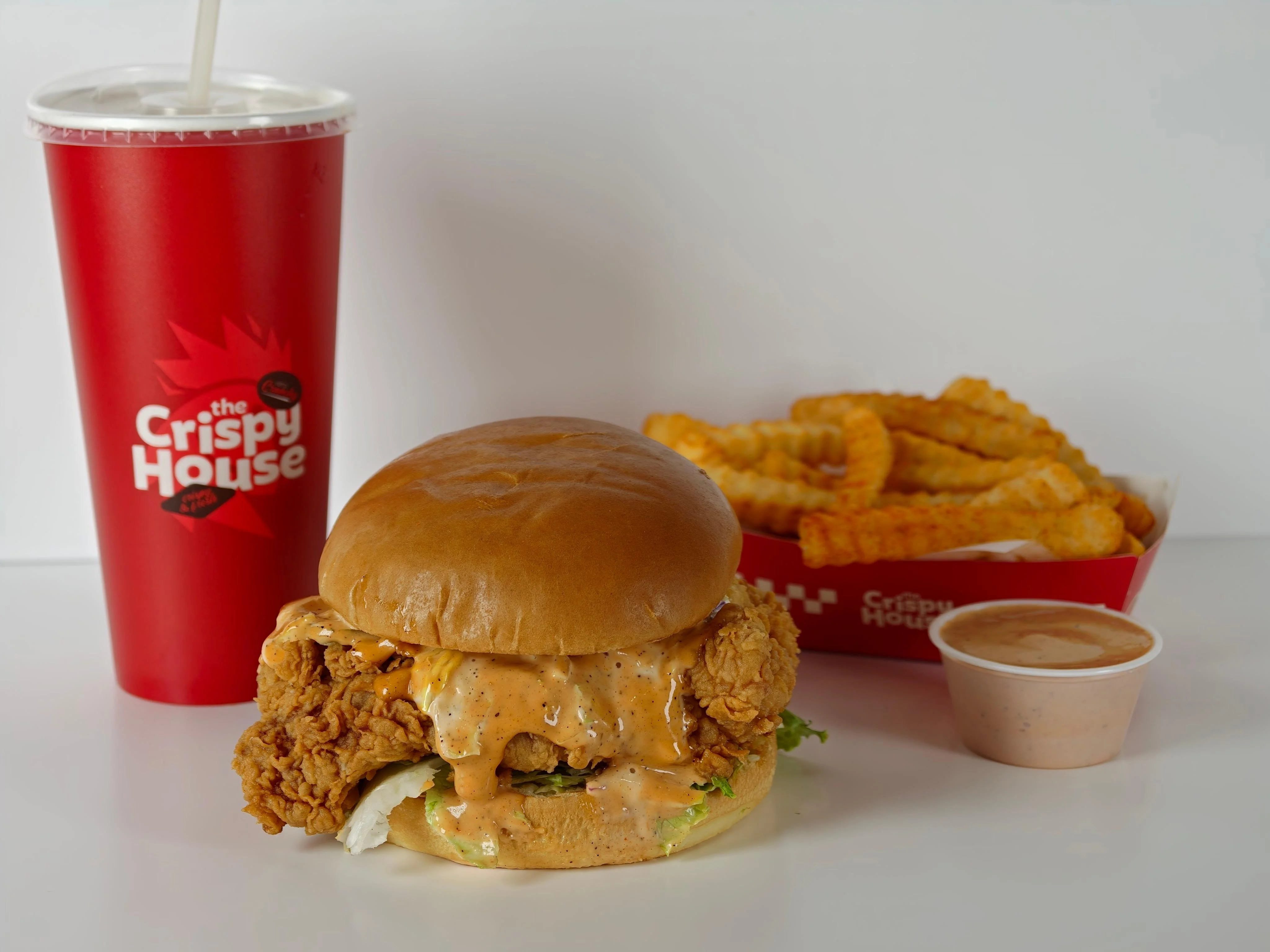 A crispy fried chicken sandwich topped with sauce, served with a side of crinkle-cut fries and a drink.
