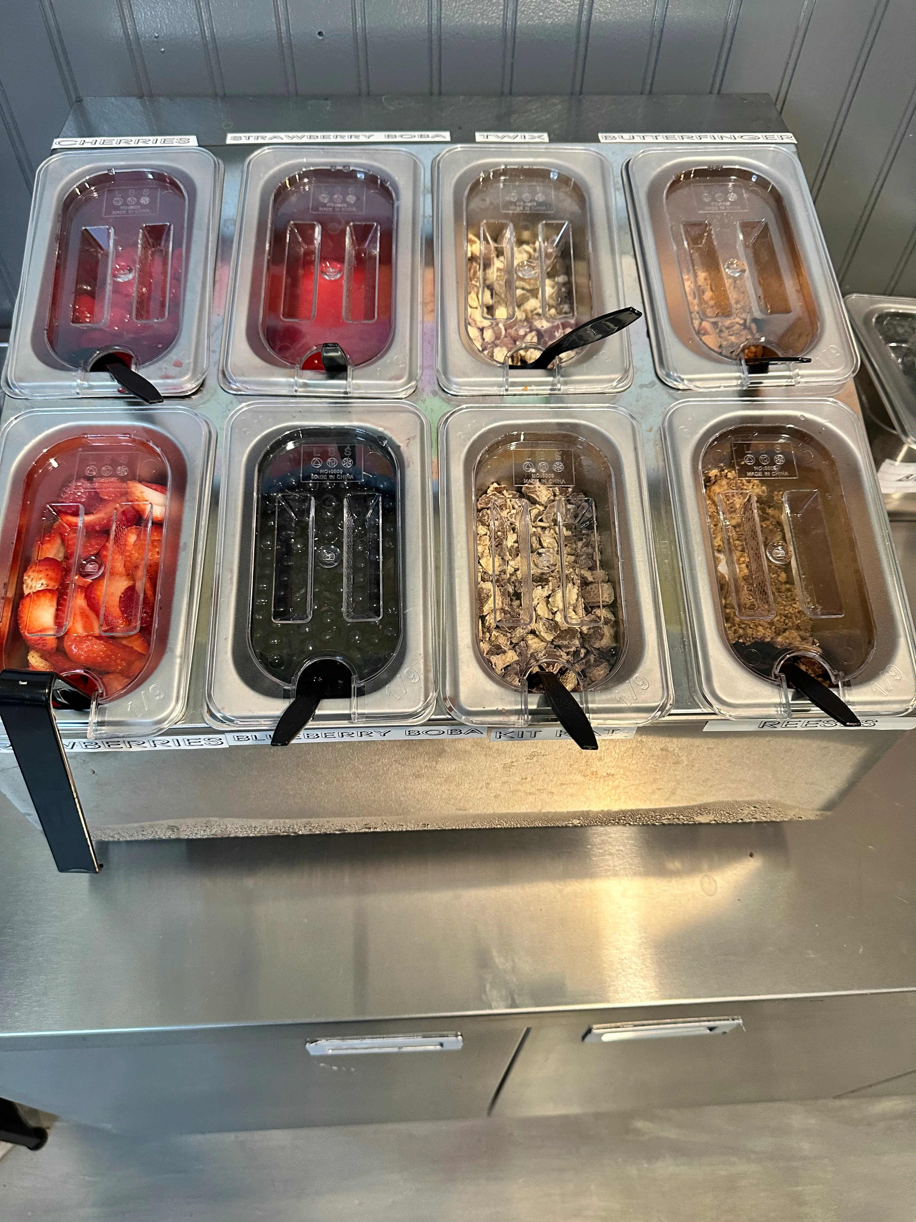 A display of various toppings in containers, including fruits, boba, and candy options for frozen yogurt.