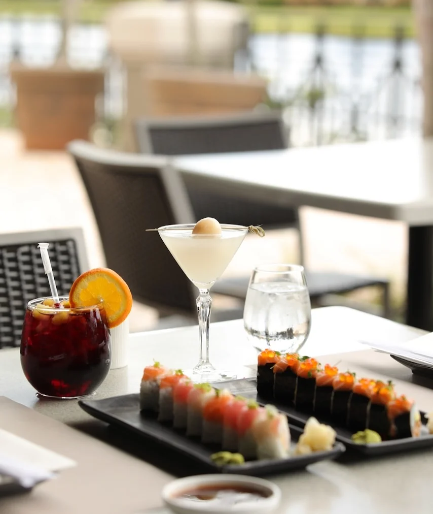 An outdoor restaurant table featuring two platters of specialty sushi rolls, a lychee martini, and a dark berry cocktail garnished with an orange slice.