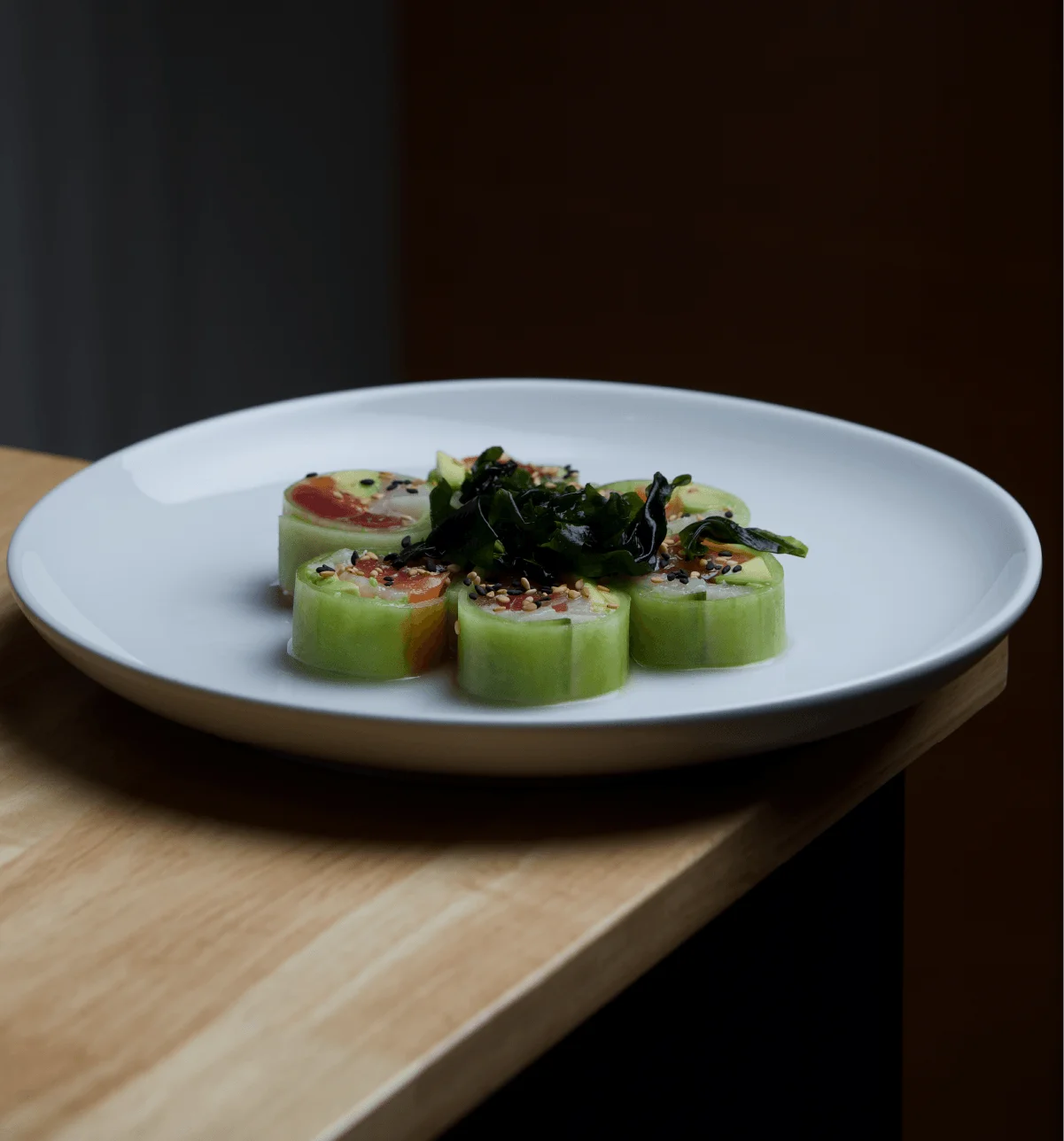 A cluster of riceless sushi rolls wrapped in thin cucumber sheets instead of seaweed, filled with tuna, salmon, and avocado, sprinkled with sesame seeds and topped with dark seaweed salad, served on a white round plate sitting on a light wooden table.
