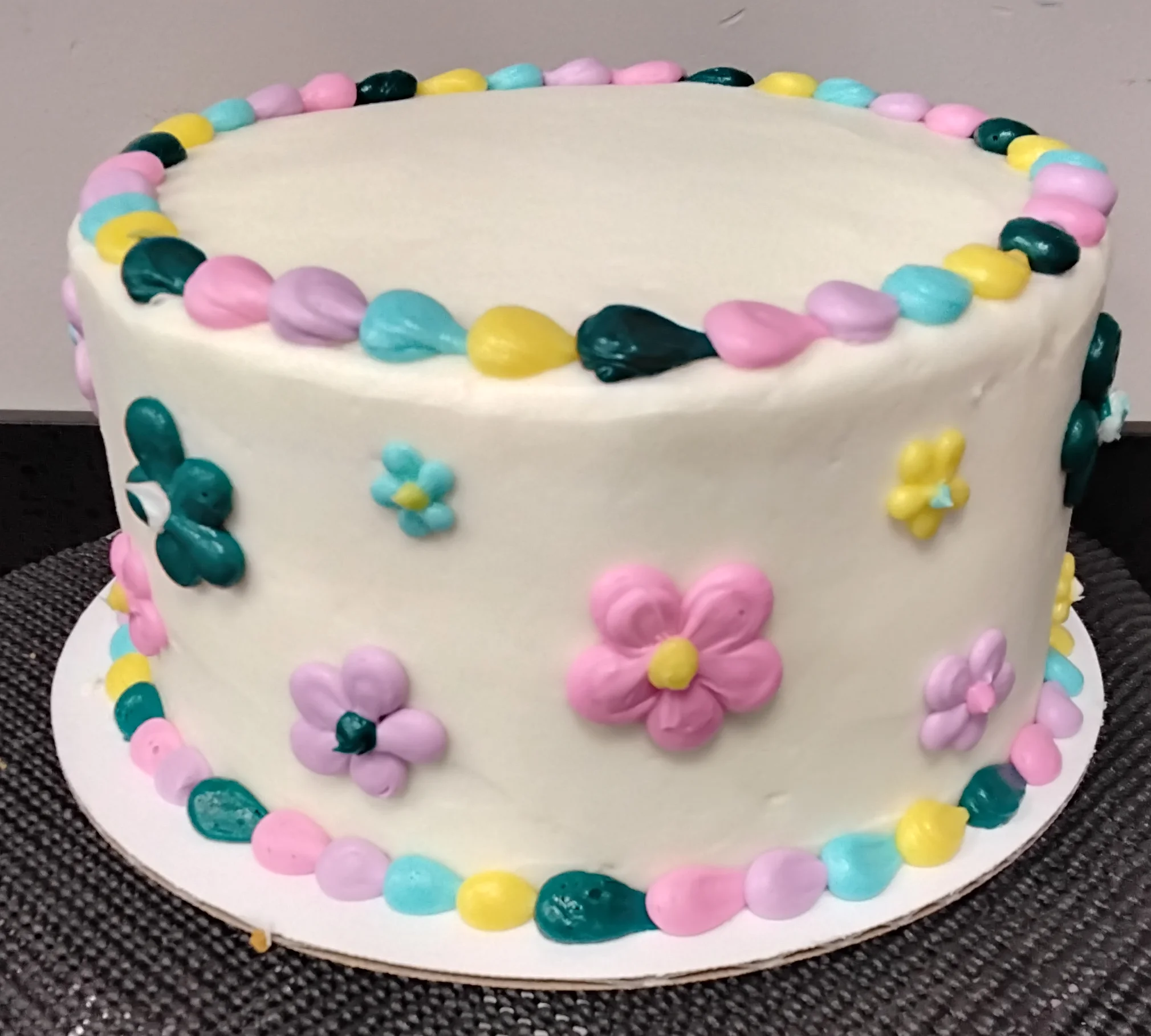 Round cake with dew drop styled borders in a variety of spring time colors with the sides of the cake covered in the same colored flowers in different sizes.