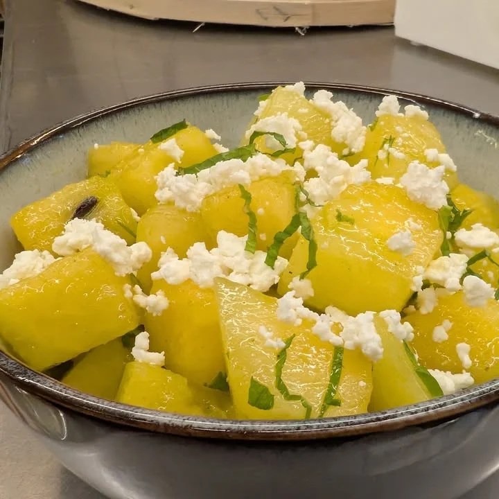 A bowl filled with diced yellow watermelon, topped with crumbled cheese and fresh mint leaves, offering a refreshing summer salad.