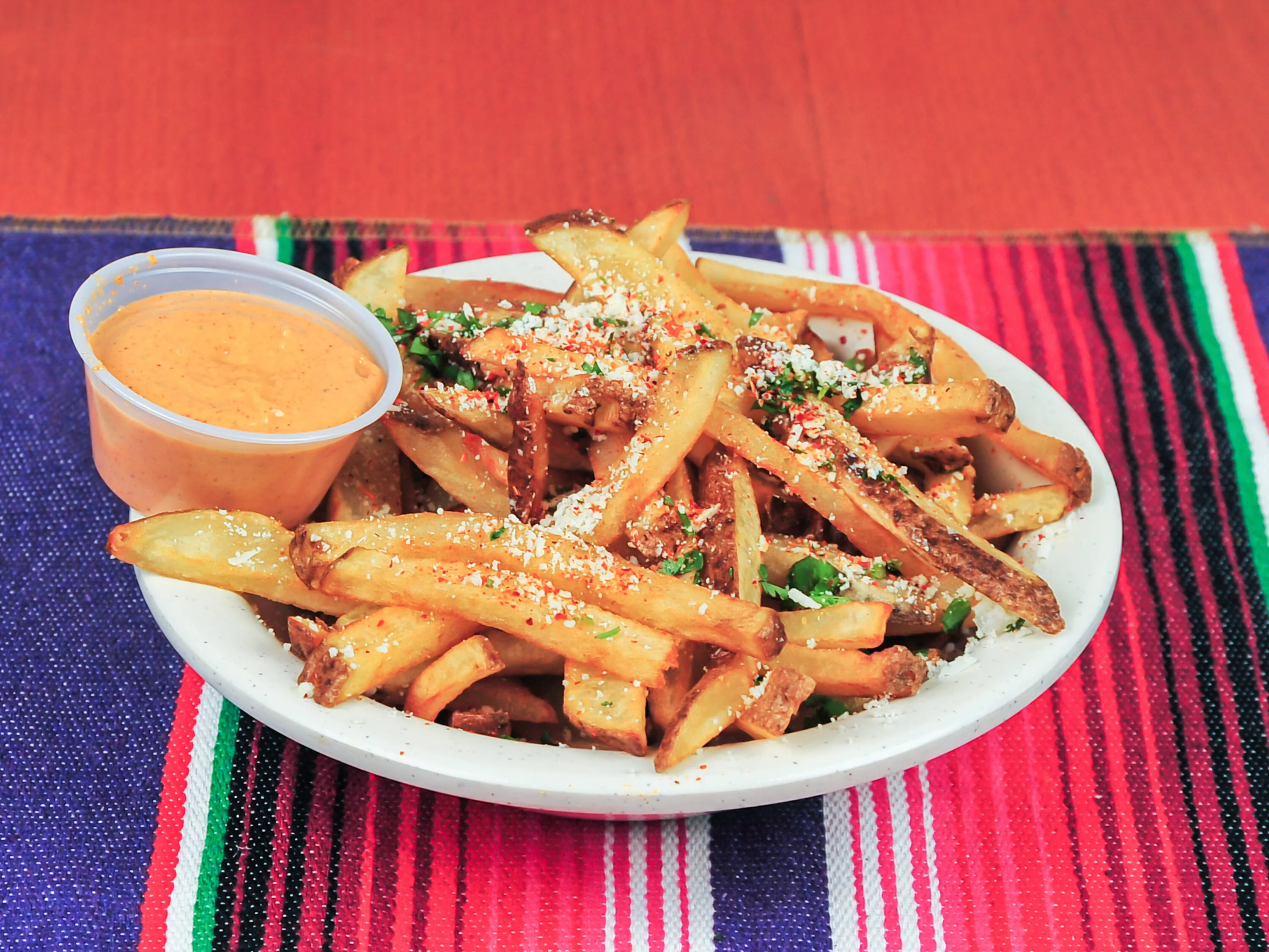 A serving of seasoned French fries topped with fresh herbs and sprinkled cheese, served with a side of creamy dipping sauce. The dish is placed on a vibrant table runner.