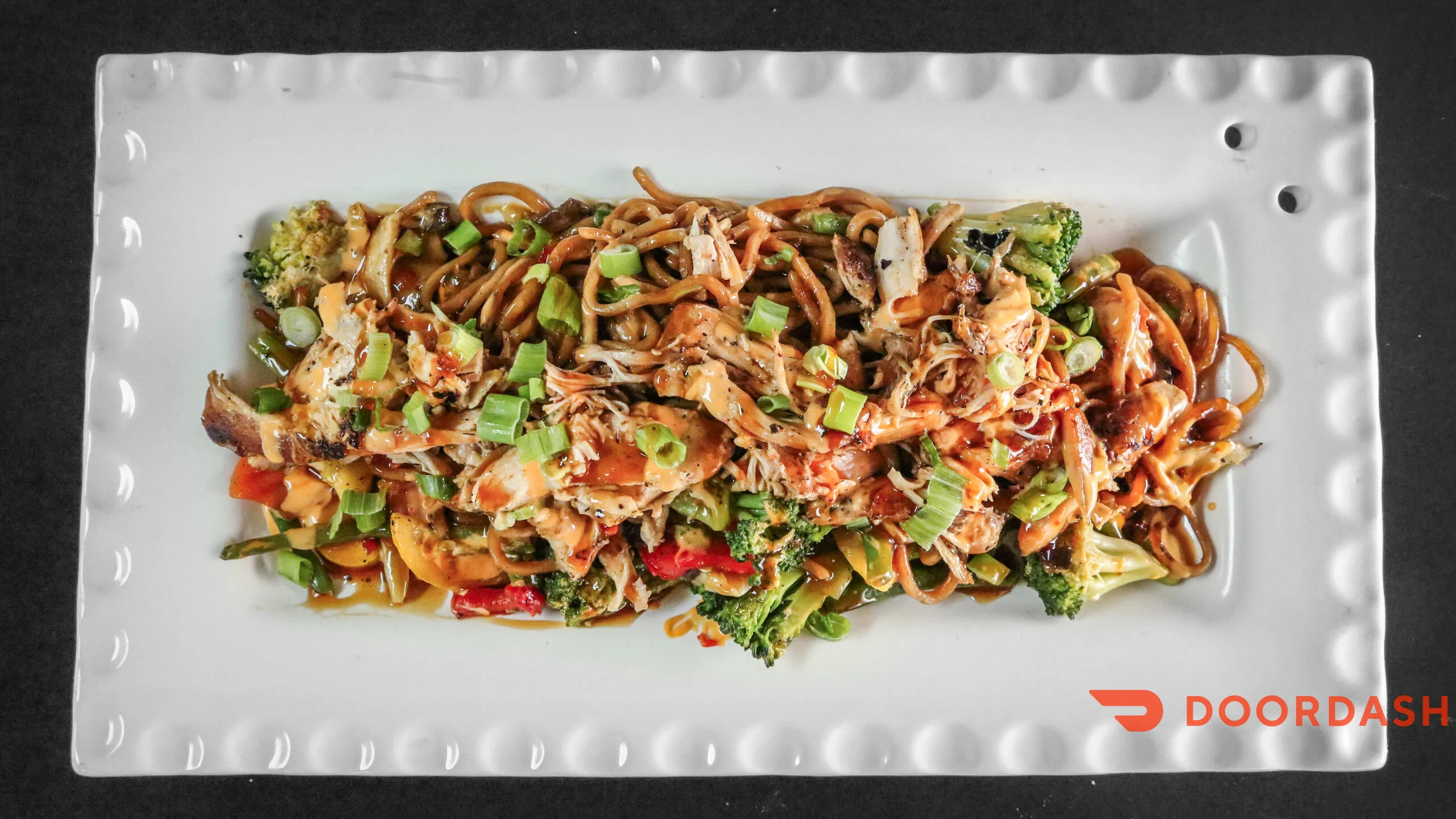 A plate of stir-fried noodles with shredded chicken and mixed vegetables, including broccoli and bell peppers. The dish is garnished with chopped green onions.