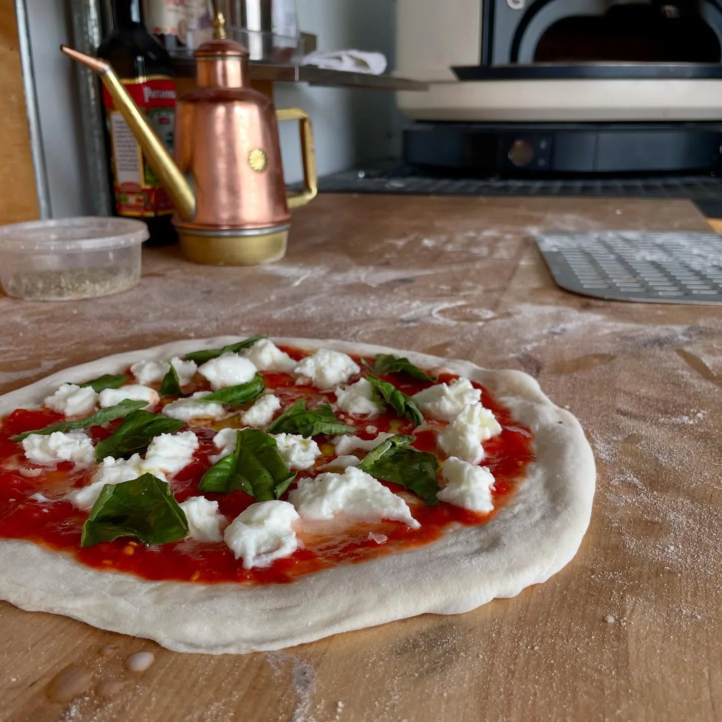 An uncooked pizza base topped with marinara sauce, fresh mozzarella, and basil leaves, lying on a floured surface with tools in the background.
