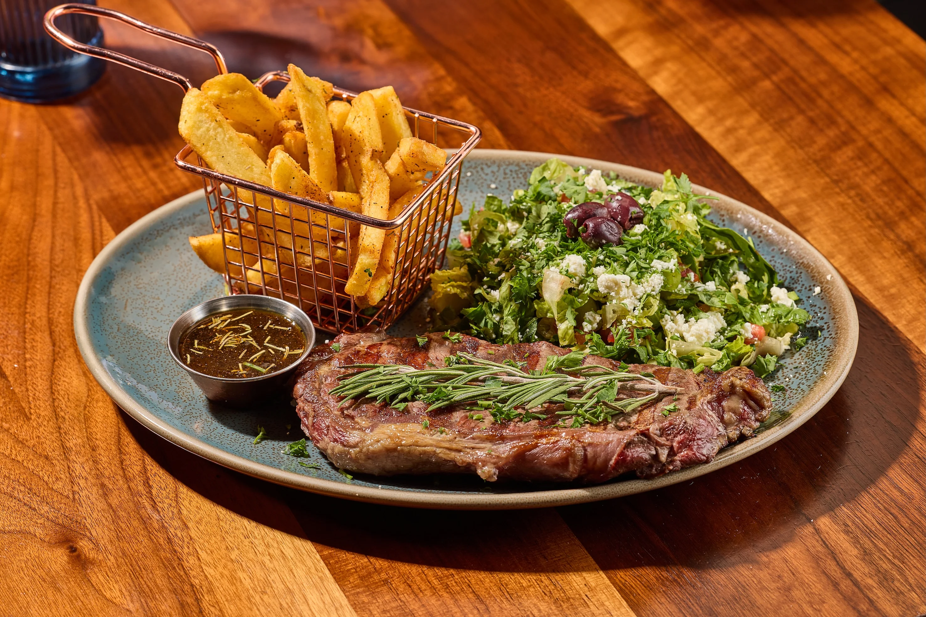 A plate with grilled steak garnished with rosemary, a side of fresh salad, and crispy fries in a basket. A small cup of sauce accompanies the steak.