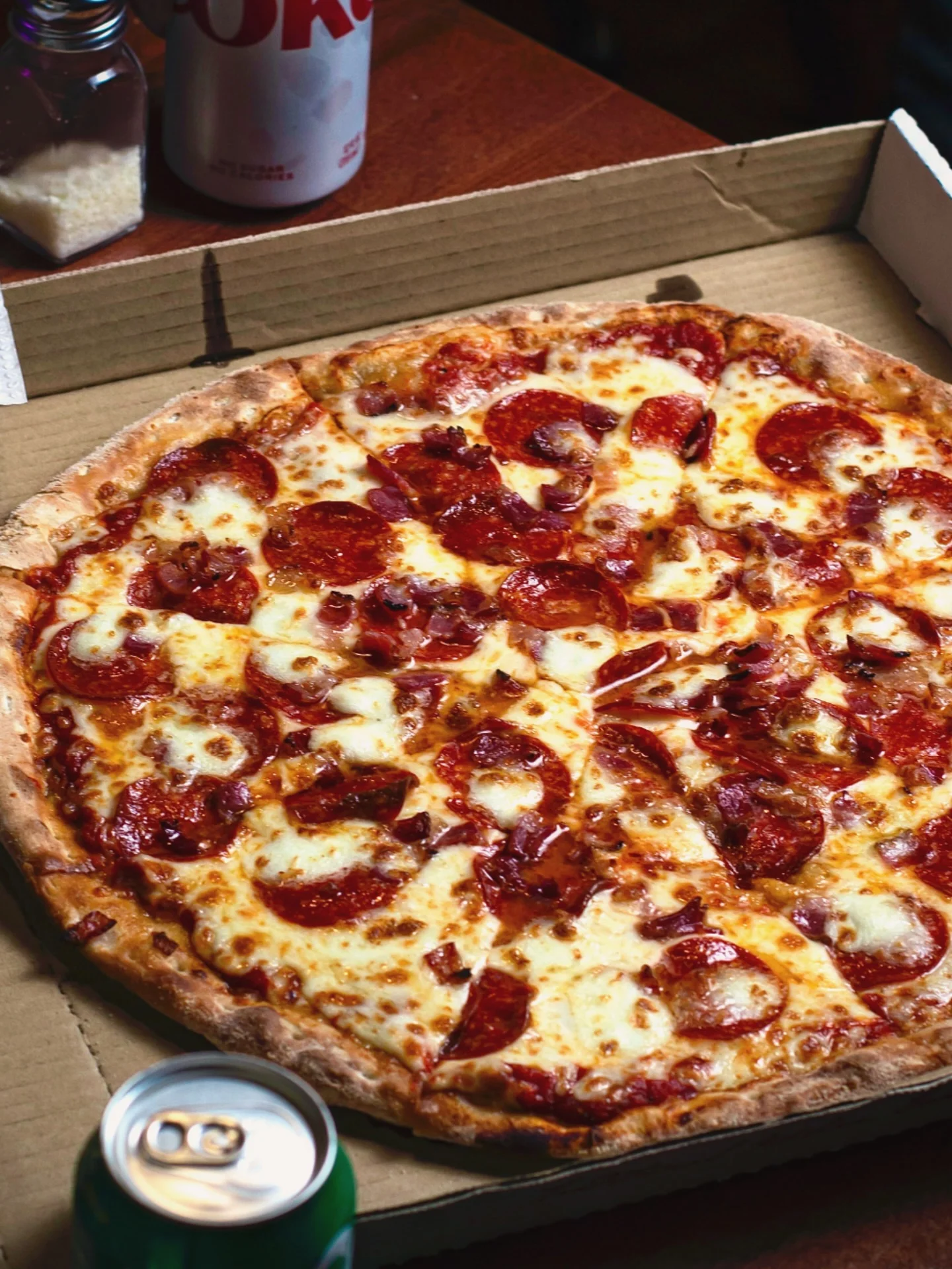 A whole pepperoni pizza in a cardboard box, with a can of soda nearby.
