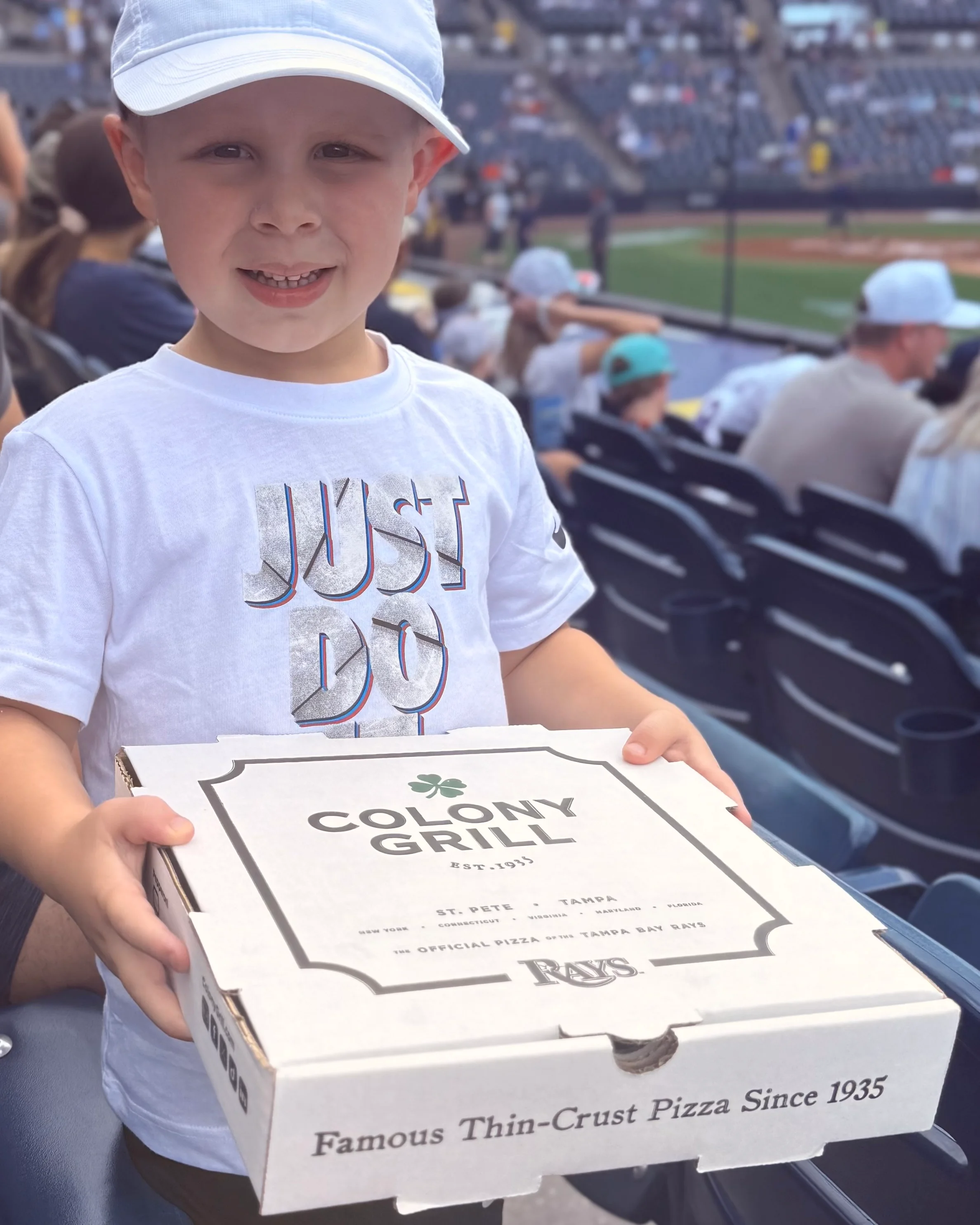 Child holding a box of Colony Grill thin-crust pizza at a baseball game