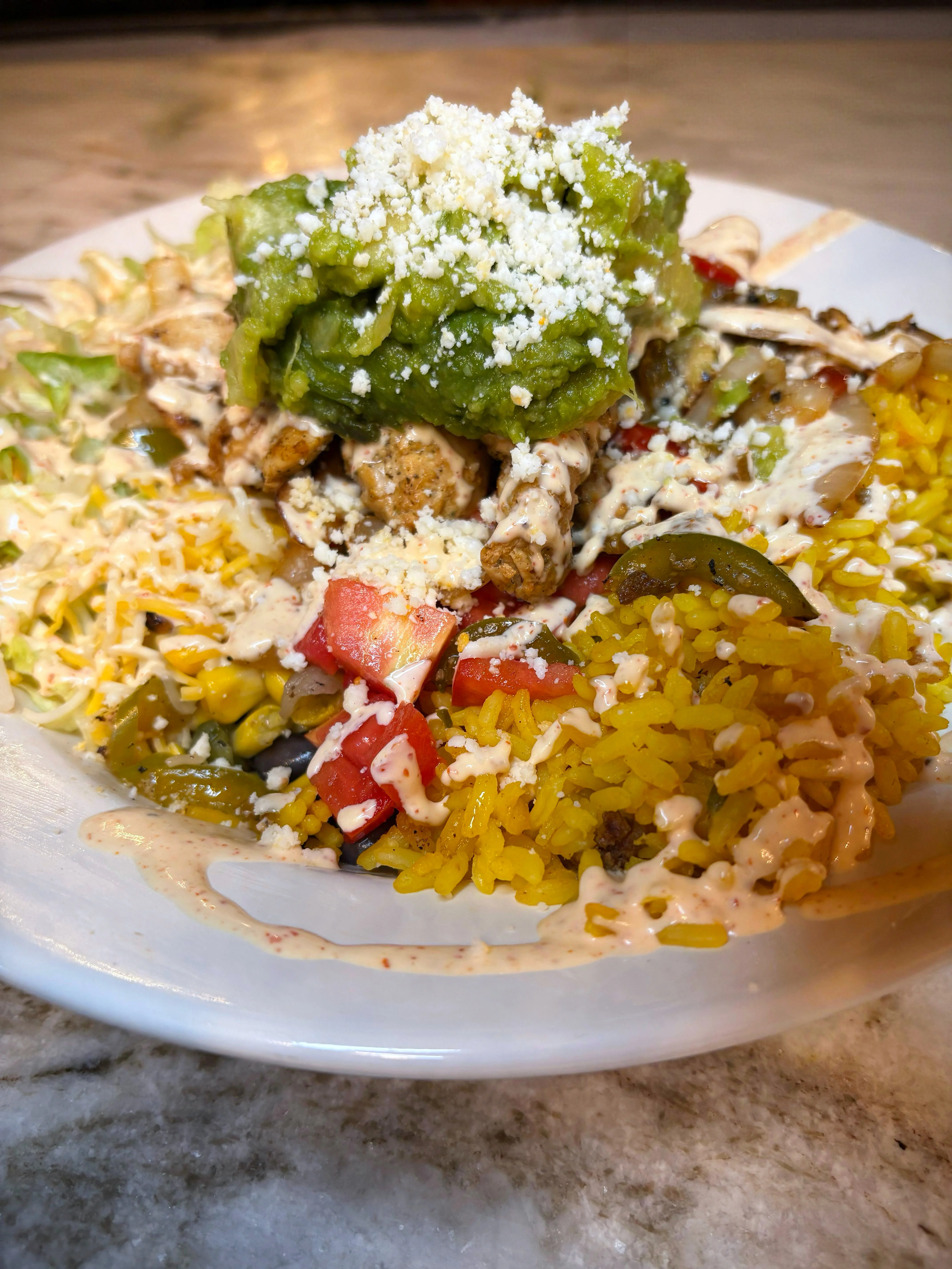 A colorful plate featuring seasoned chicken on a bed of yellow rice, garnished with guacamole and cheese. Fresh vegetables, including lettuce, tomatoes, and jalapeños are also present, creating a vibrant and appetizing dish.