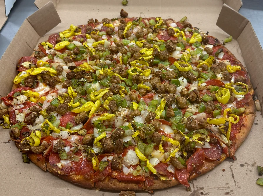 A fully loaded pizza with various toppings including pepperoni, green peppers, onions, and yellow banana peppers. The pizza is served in a cardboard box.
