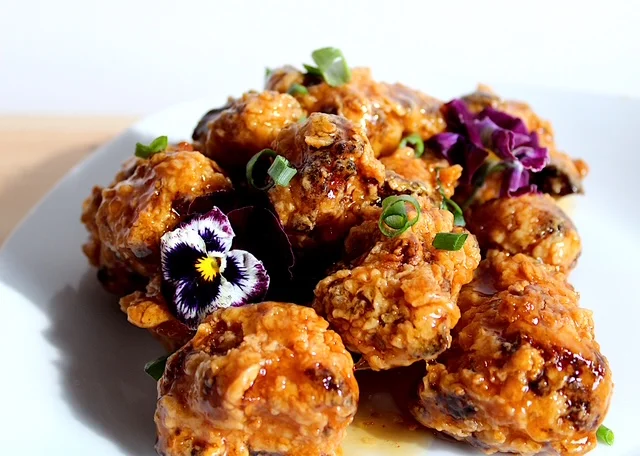 A colorful platter of crispy fried chicken bites glazed with a sweet and spicy sauce, garnished with green onions and edible flowers.